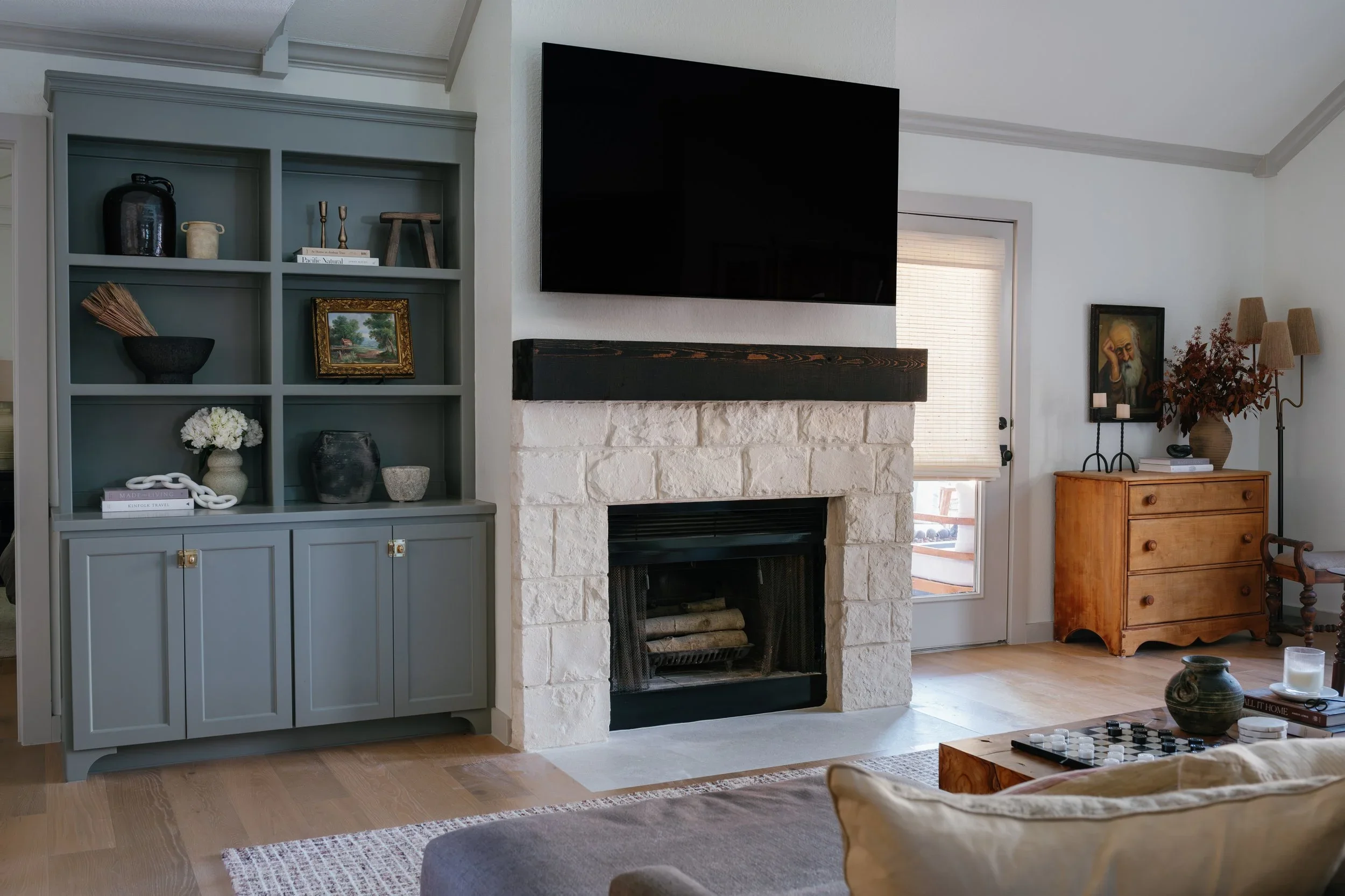 Living room with a fireplace, a wall-mounted flat-screen TV, a blue bookshelf with decorative items, a wooden side table with a framed painting, a lamp, and a vase with dried flowers. There are windows with blinds and various decorative objects on fu