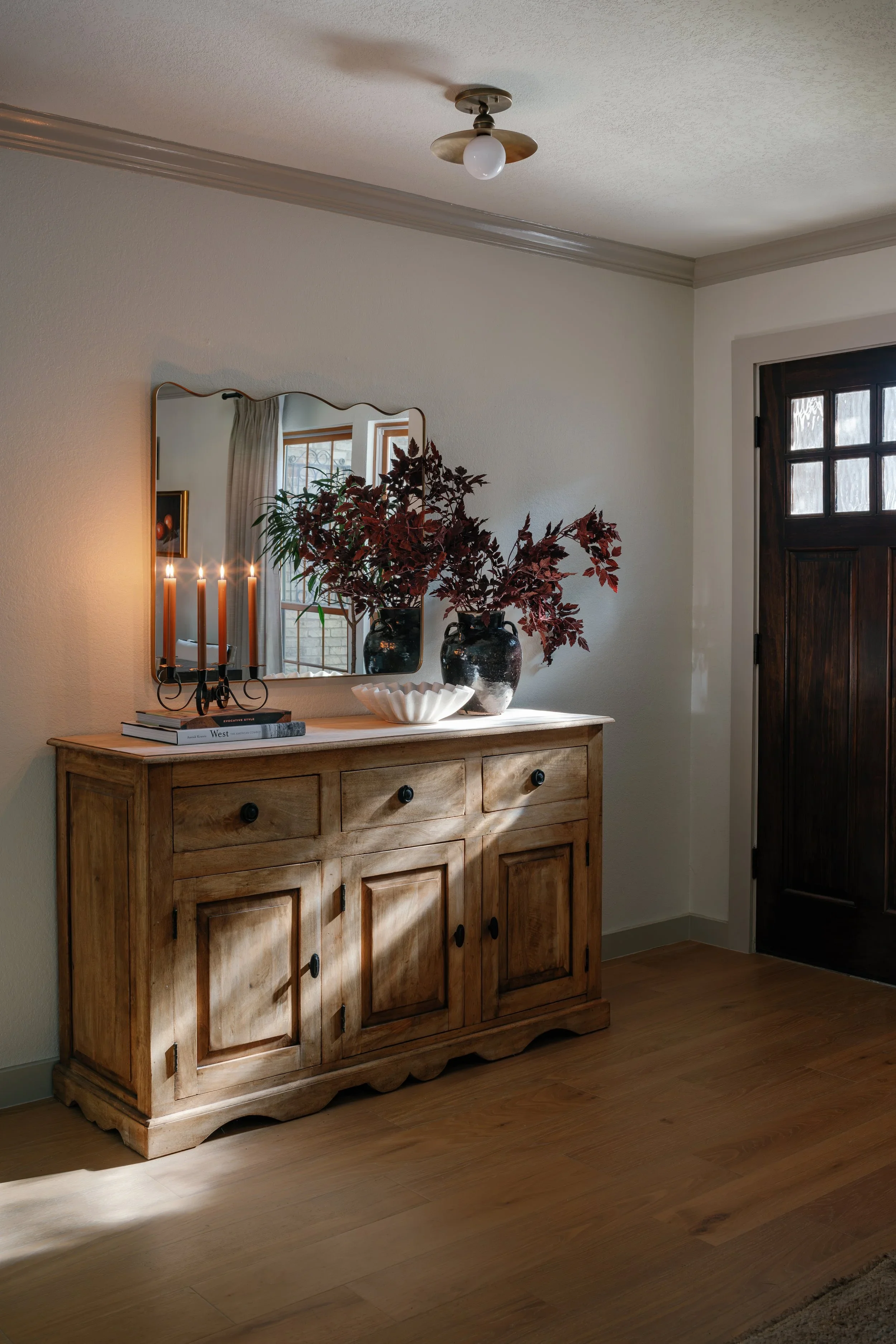 A wooden sideboard with a mirror above it, decorated with two black vases containing red leaves, a white bowl, and a pair of books, inside a room with a window and a dark wooden door.