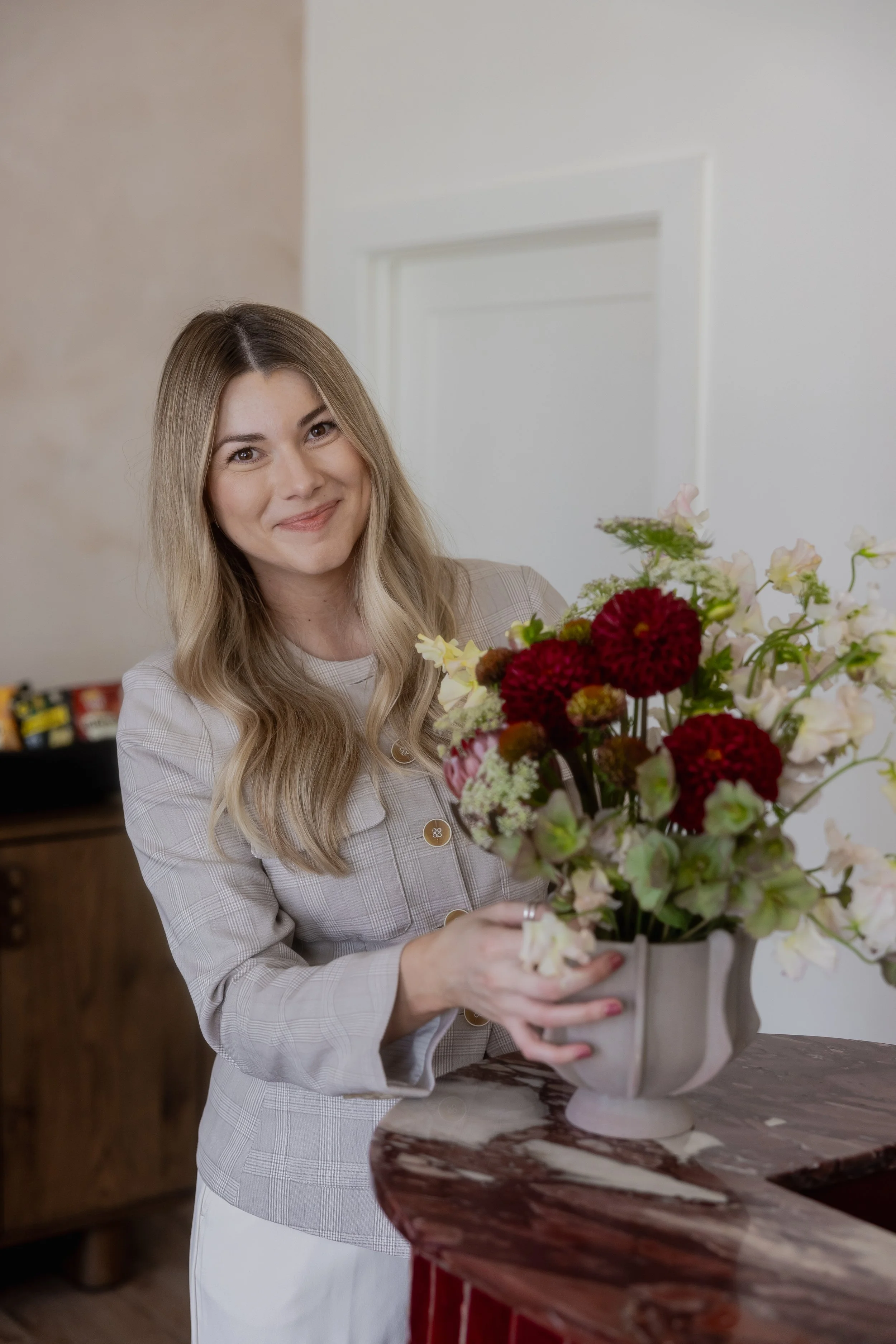 A woman with long blonde hair holding a large bouquet of mixed flowers, smiling, in an indoor setting.