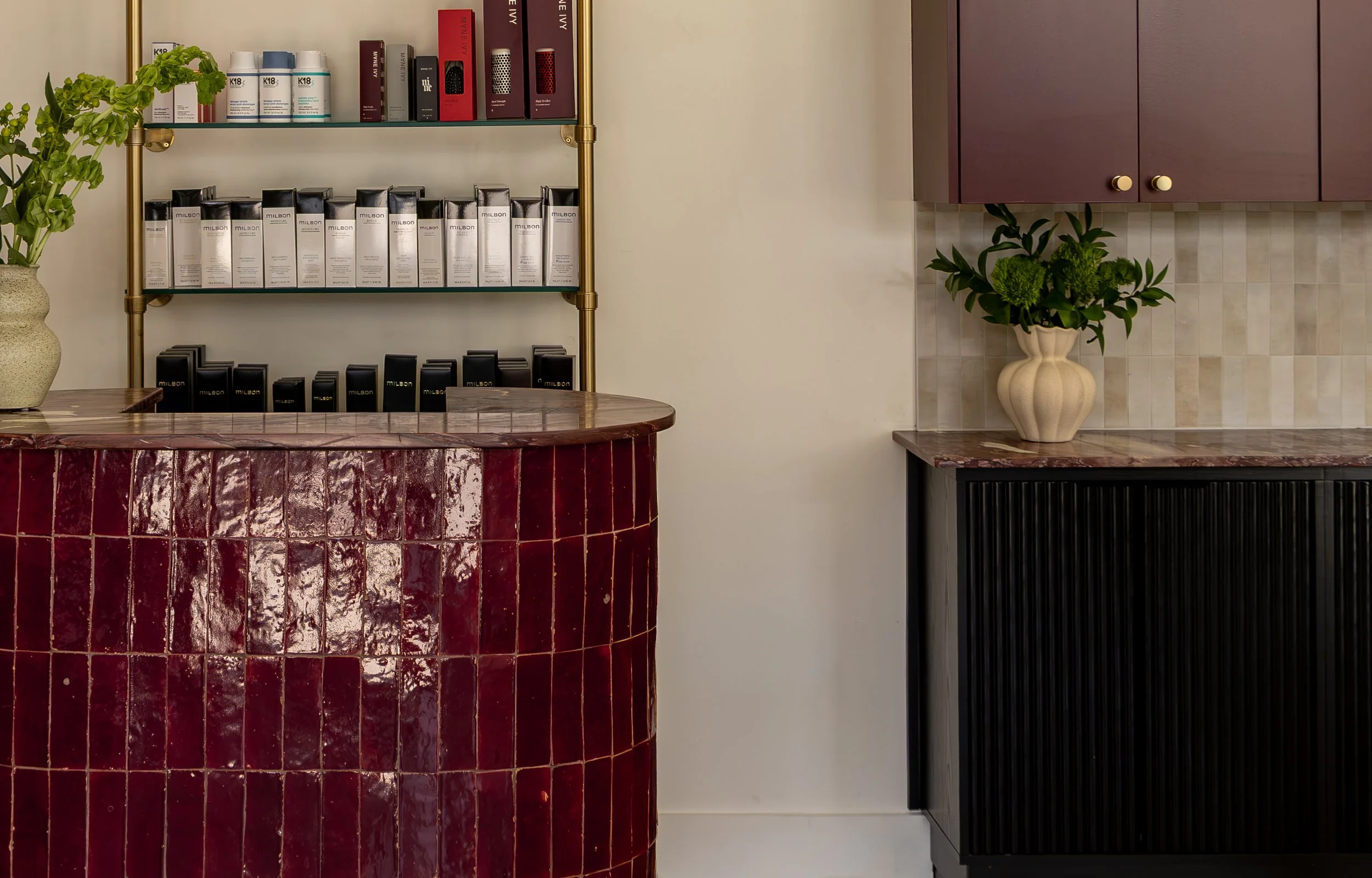 A reception area with a curved, glossy red tiled countertop, a glass shelf with hair care products, and a purple cabinet with plant in a decorative vase.