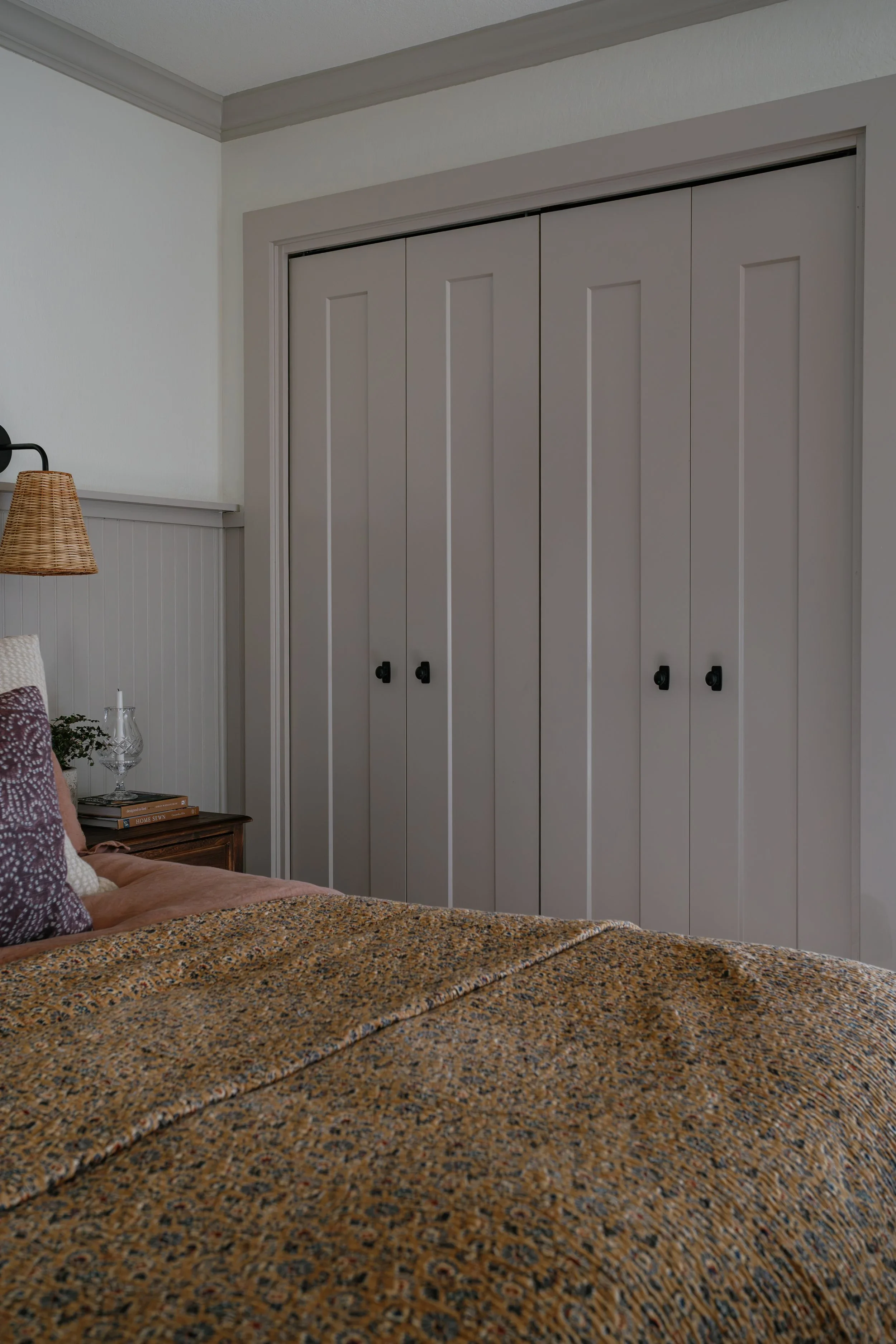 A bedroom with a bed covered in a multicolored patterned quilt, a wooden nightstand with books, a glass, and a decorative plant, a wall-mounted black lamp, white paneled beadboard wainscoting, a tan woven lampshade, and a closed closet with gray door