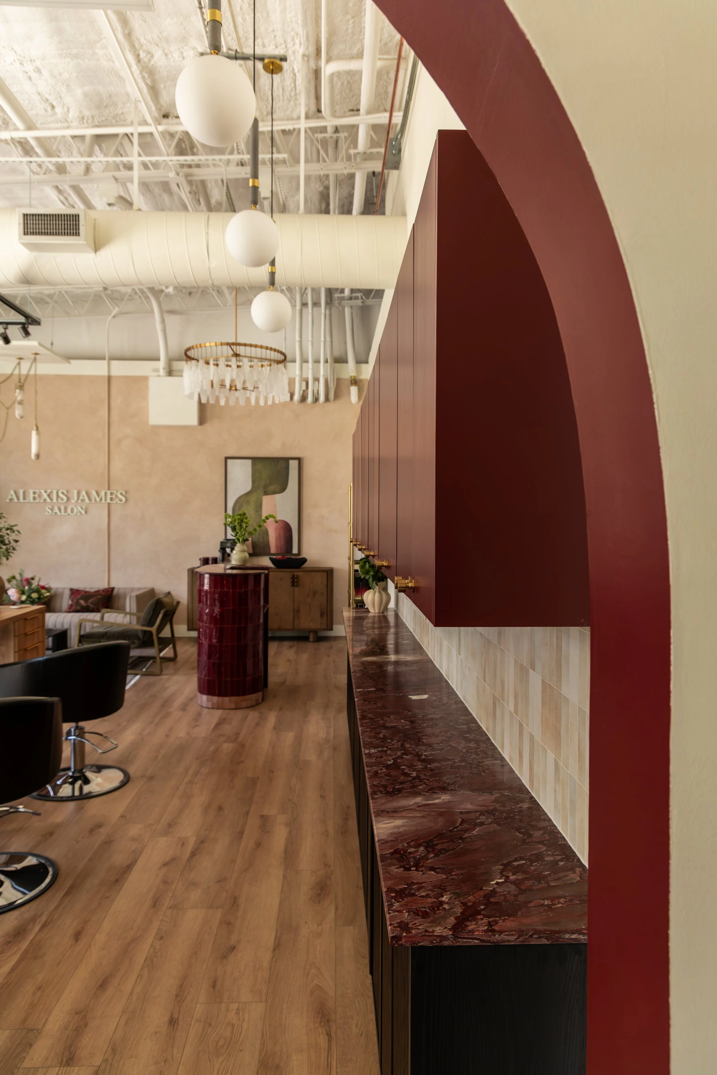 Interior of a salon with a wooden floor, modern lighting fixtures, a red marble countertop, and seating area with sofas and chairs.