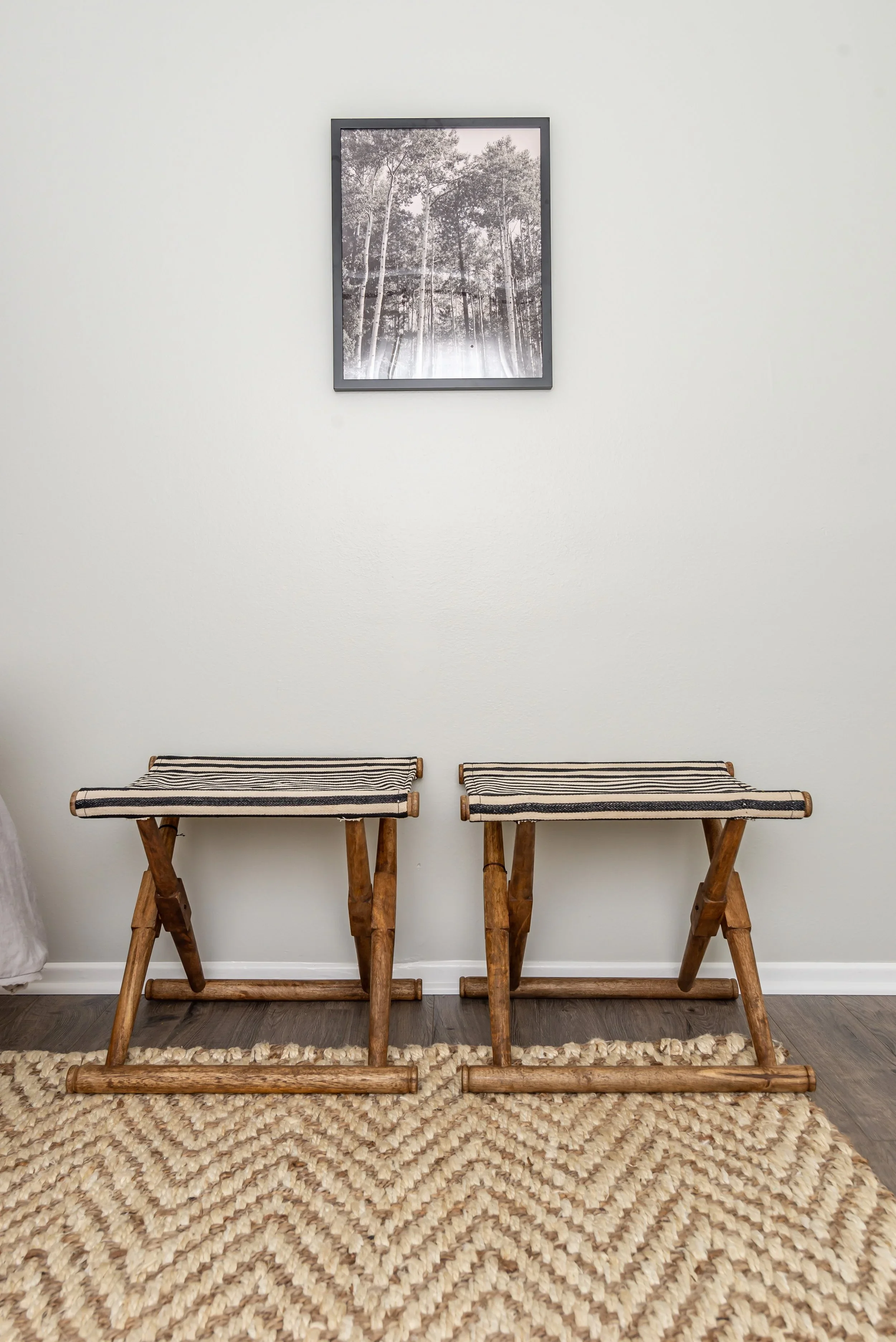 Two wooden stools with woven striped fabric seats are placed on a woven rug, against a plain white wall with a black and white framed photo of trees hanging above.