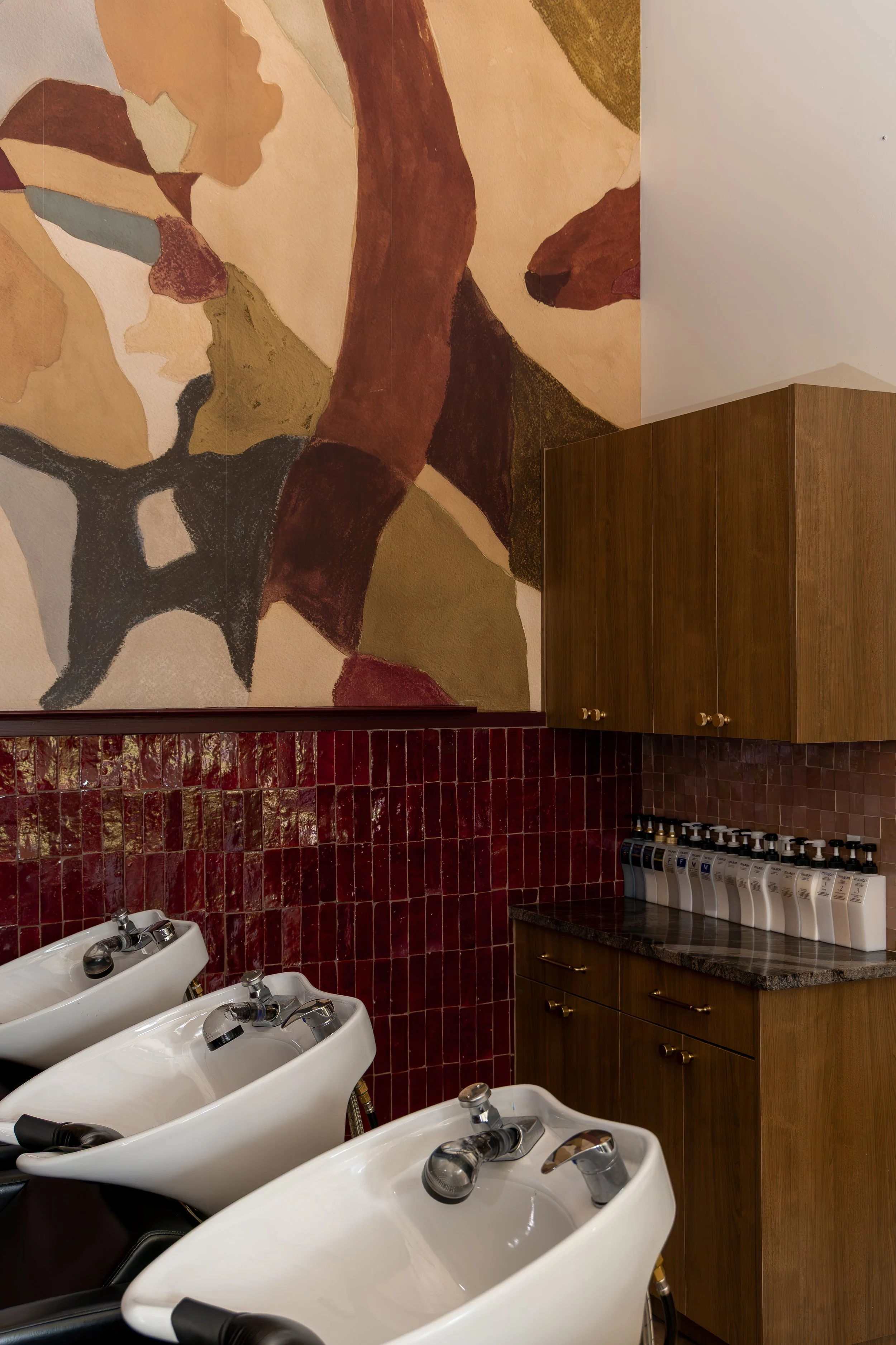 A salon or barbershop interior featuring three white hair wash sinks with black chairs, a wooden cabinet, and a wall painting with abstract earth-tone shapes. There are bottles of hair care products on a countertop.