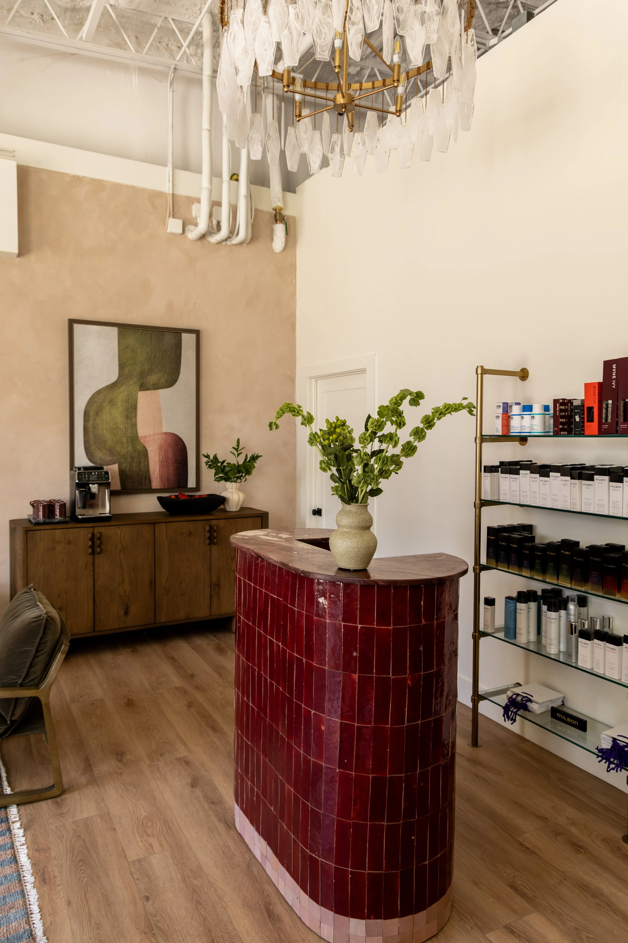 Interior view of a room with a red tiled bar or counter, a vase with green plants on it, a wooden sideboard with art, a small table with a chair, and white walls decorated with artwork. There is a chandelier hanging from the ceiling and a shelf with 