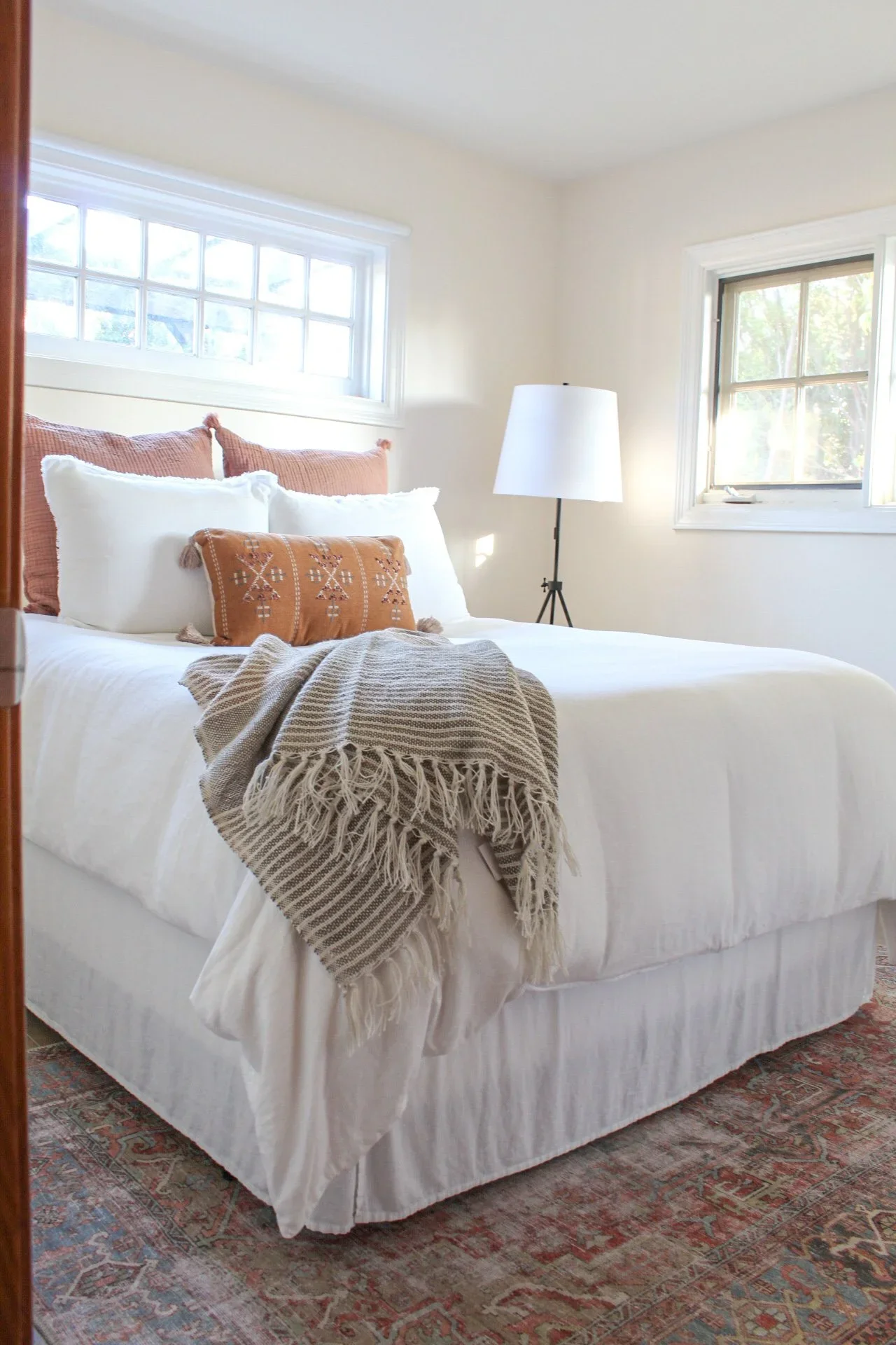 A neatly made bed with white linens, beige decorative pillows, a fringed beige throw blanket, and a patterned area rug beneath. Two windows and a floor lamp are visible in the background.