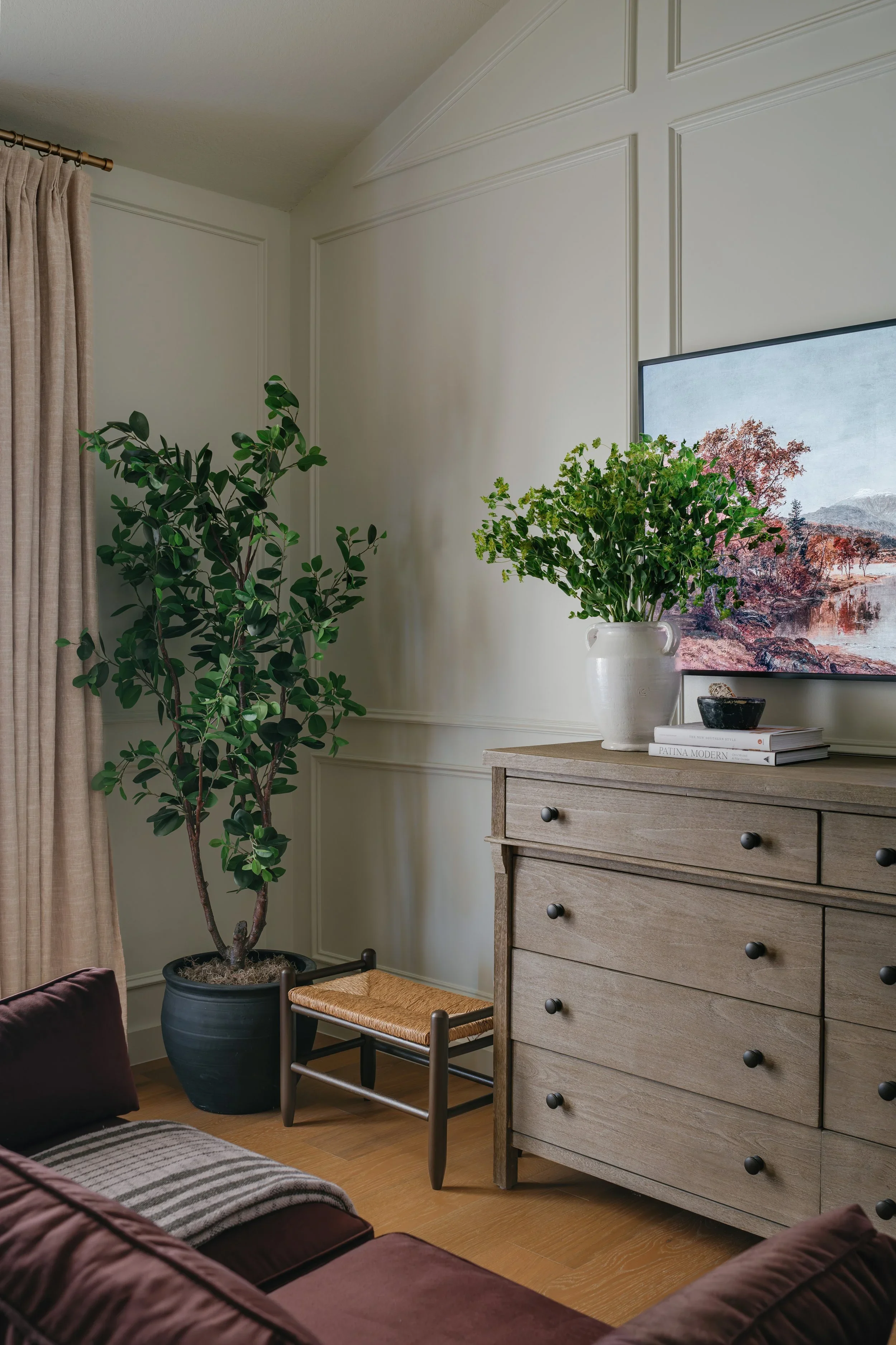 Corner of a living room with a tall potted green plant, a wooden dresser with black knobs, a white vase with green foliage on top, a television, and a beige curtain.