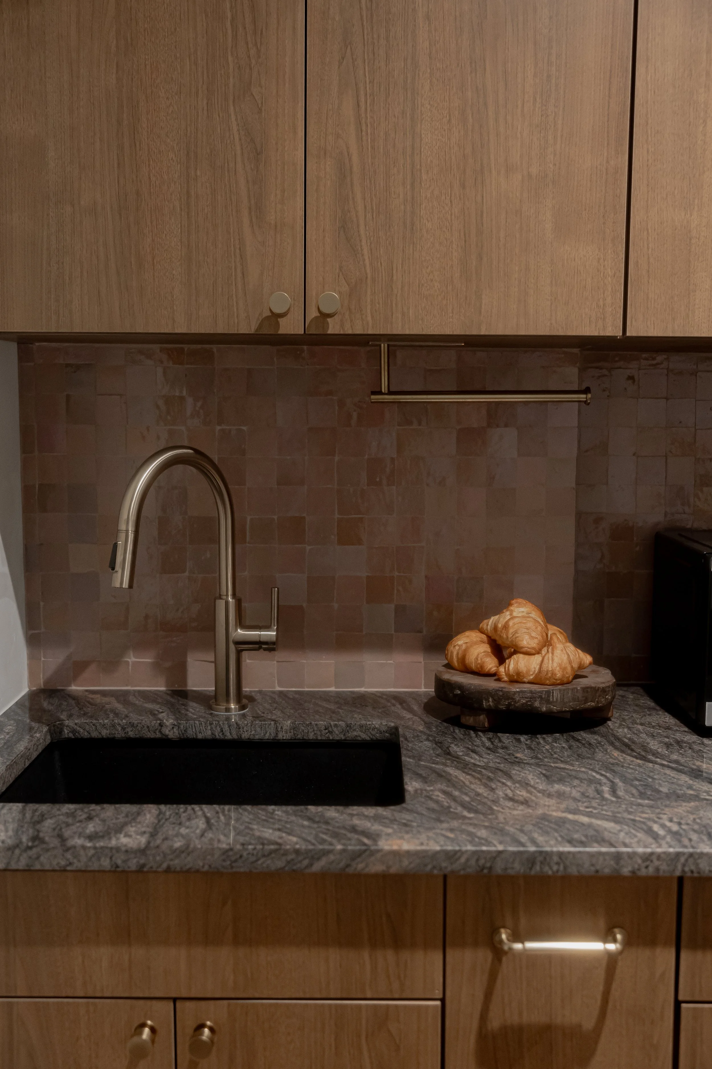 Kitchen with wooden cabinets, a marble countertop, a sink with a modern faucet, a plate of croissants, and a beige tiled backsplash.