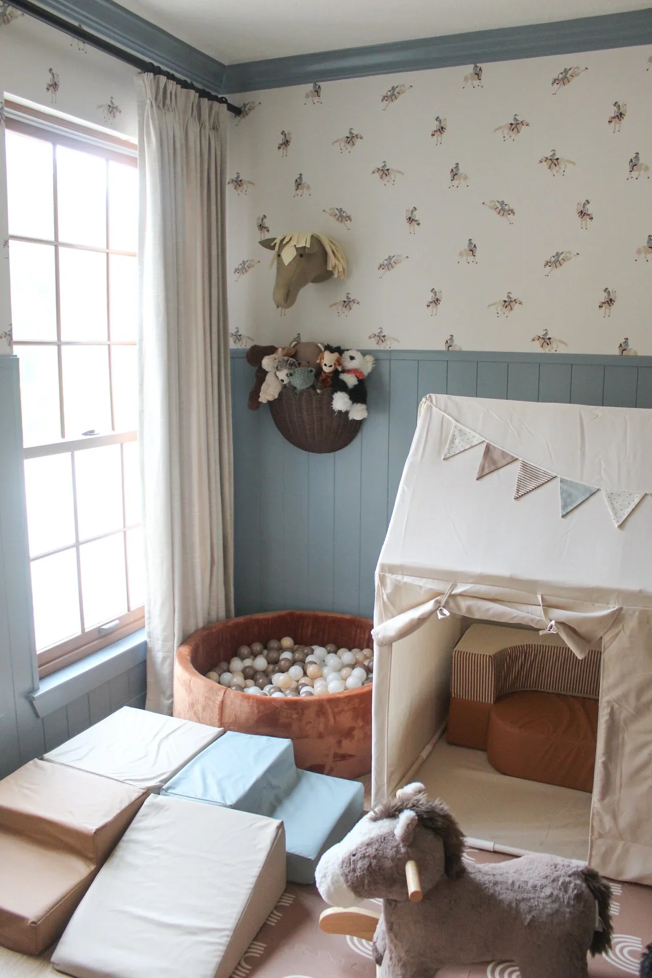 A cozy nursery with a window and curtain on the left, horse-themed wallpaper, plush toys on a wall basket, and a ball pit with white balls. There is a small tent and plush rocker on the right.