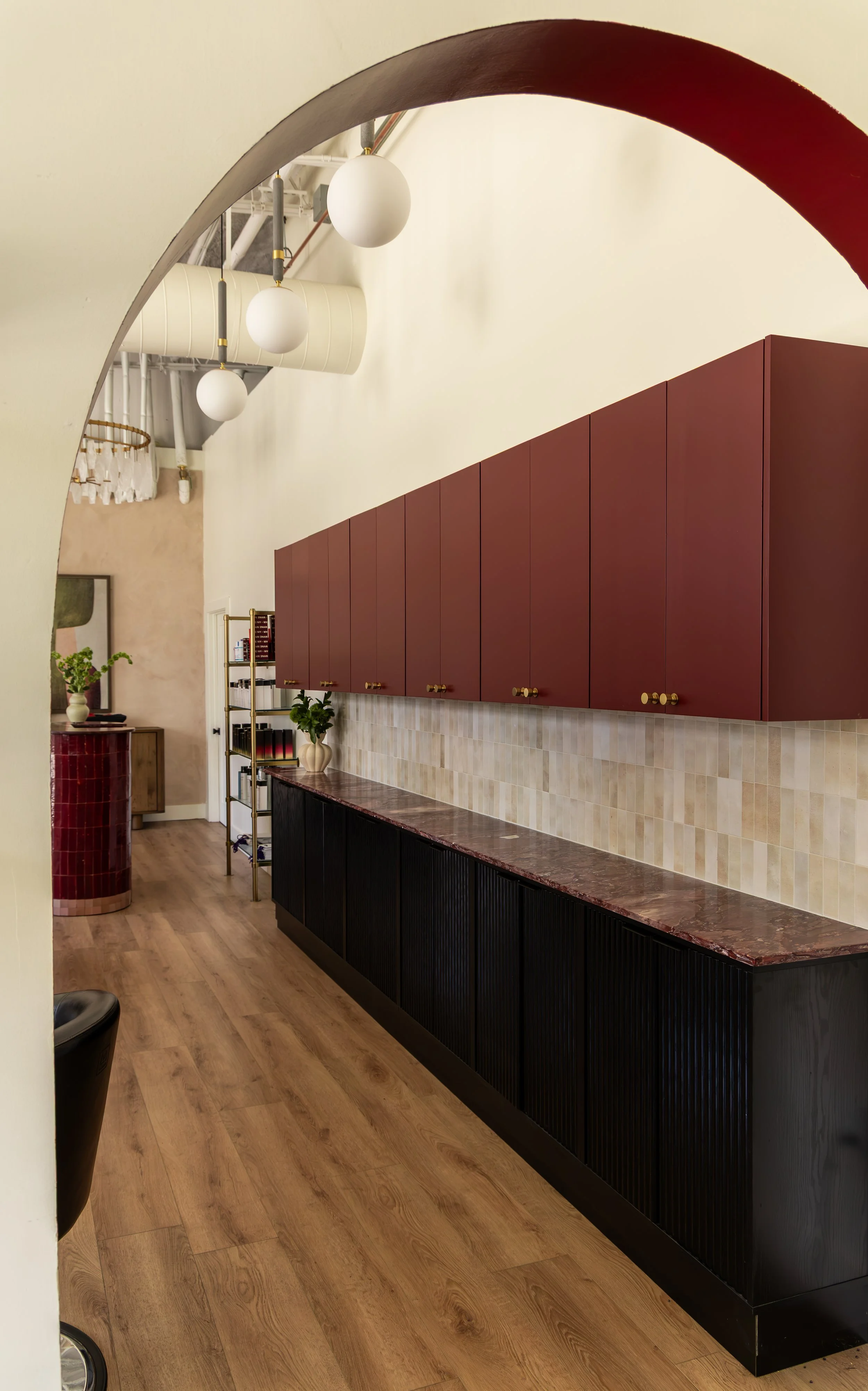 Interior of an office or lounge area with burgundy wall cabinets, black lower cabinets with vertical ridges, a marble countertop, wood flooring, and decorative items including plants and a mirror.
