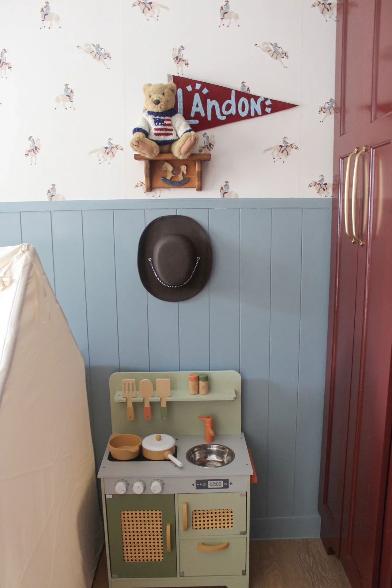 A small children's play kitchen set with a stovetop, oven, and a sink, decorated with small utensils and cookware. A teddy bear wearing a sweater is sitting on a shelf above, and a cowboy hat is hanging on the wall behind it. There is a wall with wal