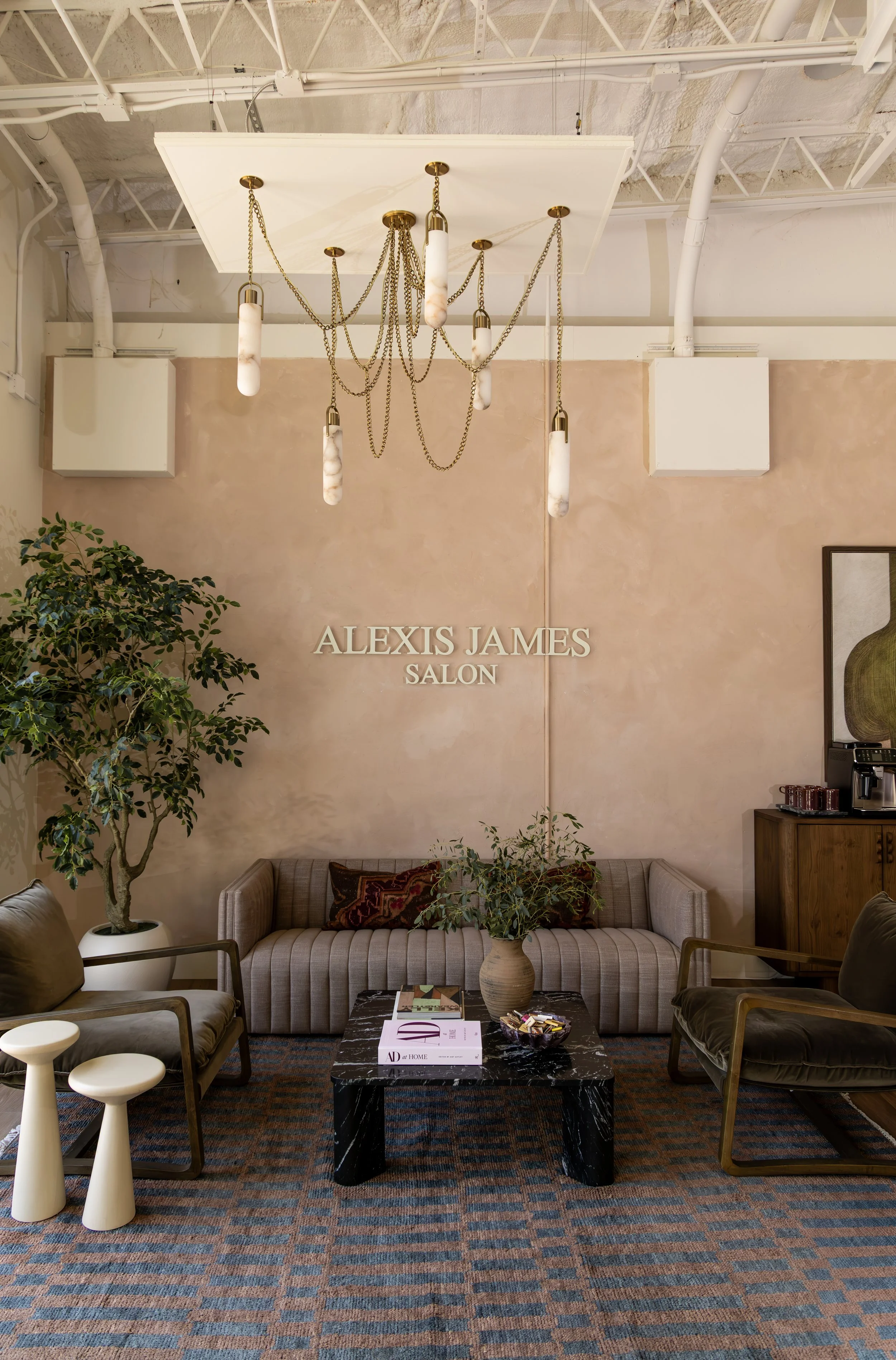 An interior lounge with a beige wall and patterned carpet. There is a striped sofa with decorative pillows, a side table, a large potted plant, and a black marble coffee table with magazines and candy. A chandelier with hanging chains and marble-like pendant lights hangs from the ceiling. The salon sign says "ALEXIS JAMES SALON."
