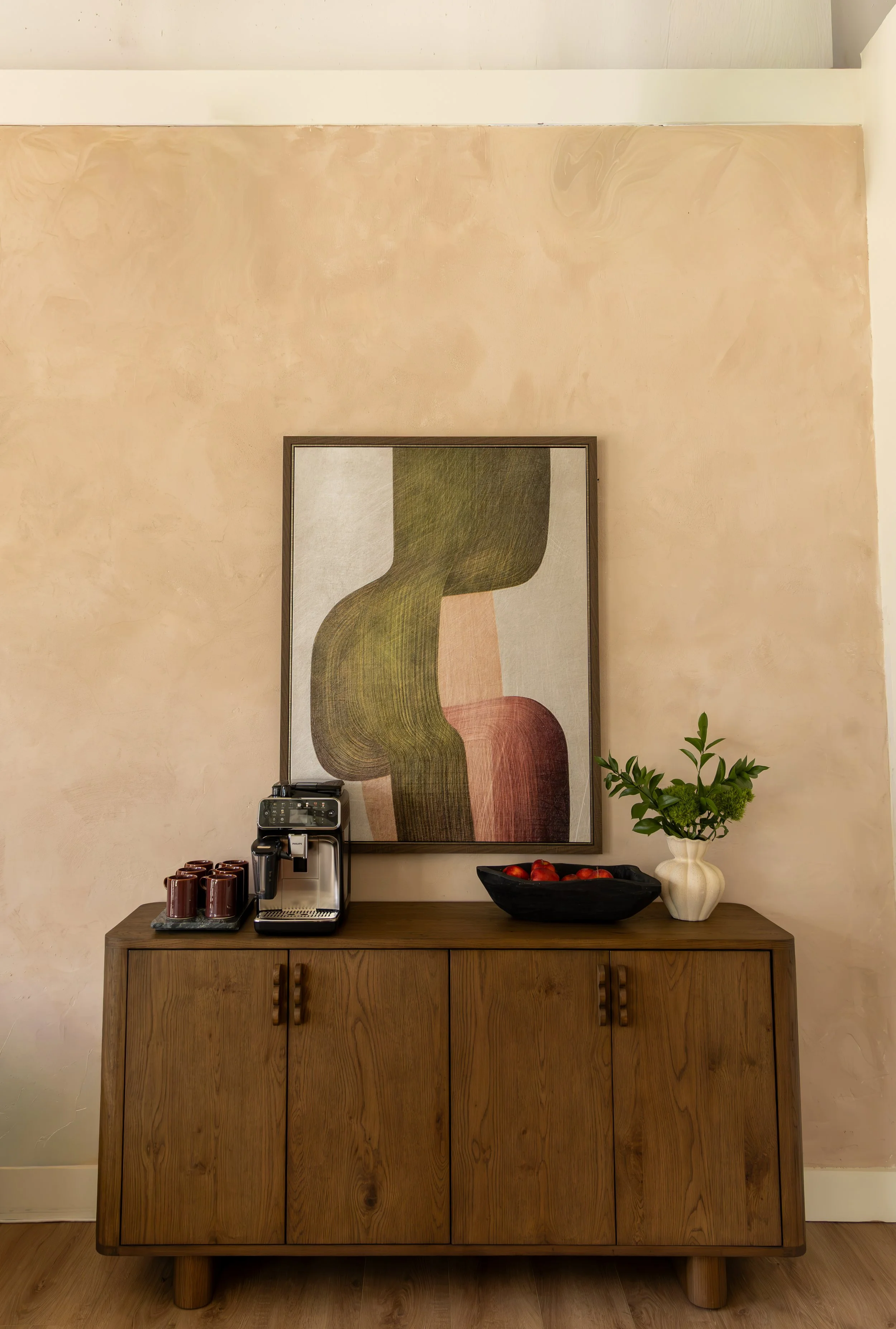 A wooden sideboard with a coffee machine, a bowl of tomatoes, and a plant with green leaves and small white flowers, in front of a beige wall with abstract art.