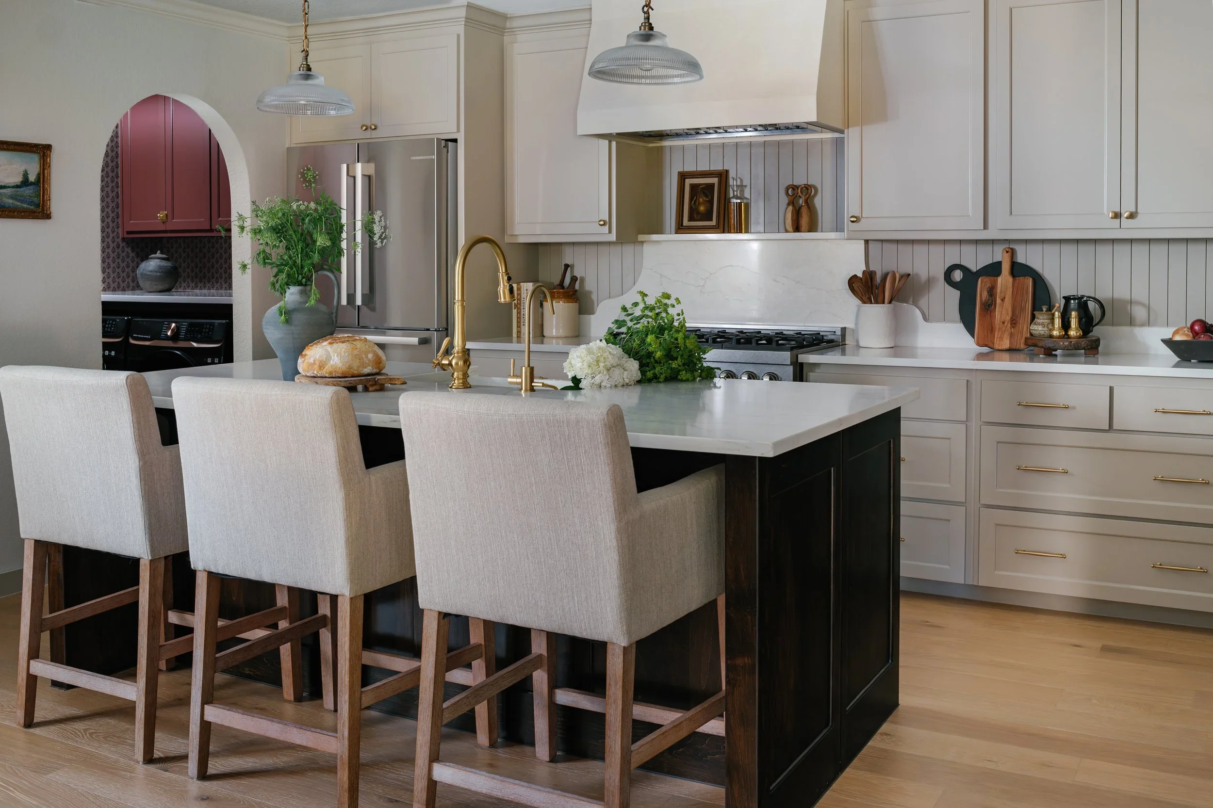 A modern kitchen with bar stool seating at a large island, featuring a gold faucet, fresh herbs, bread, and decorative items on the counter. The background includes white cabinetry, a stainless steel refrigerator, and pendant lighting.
