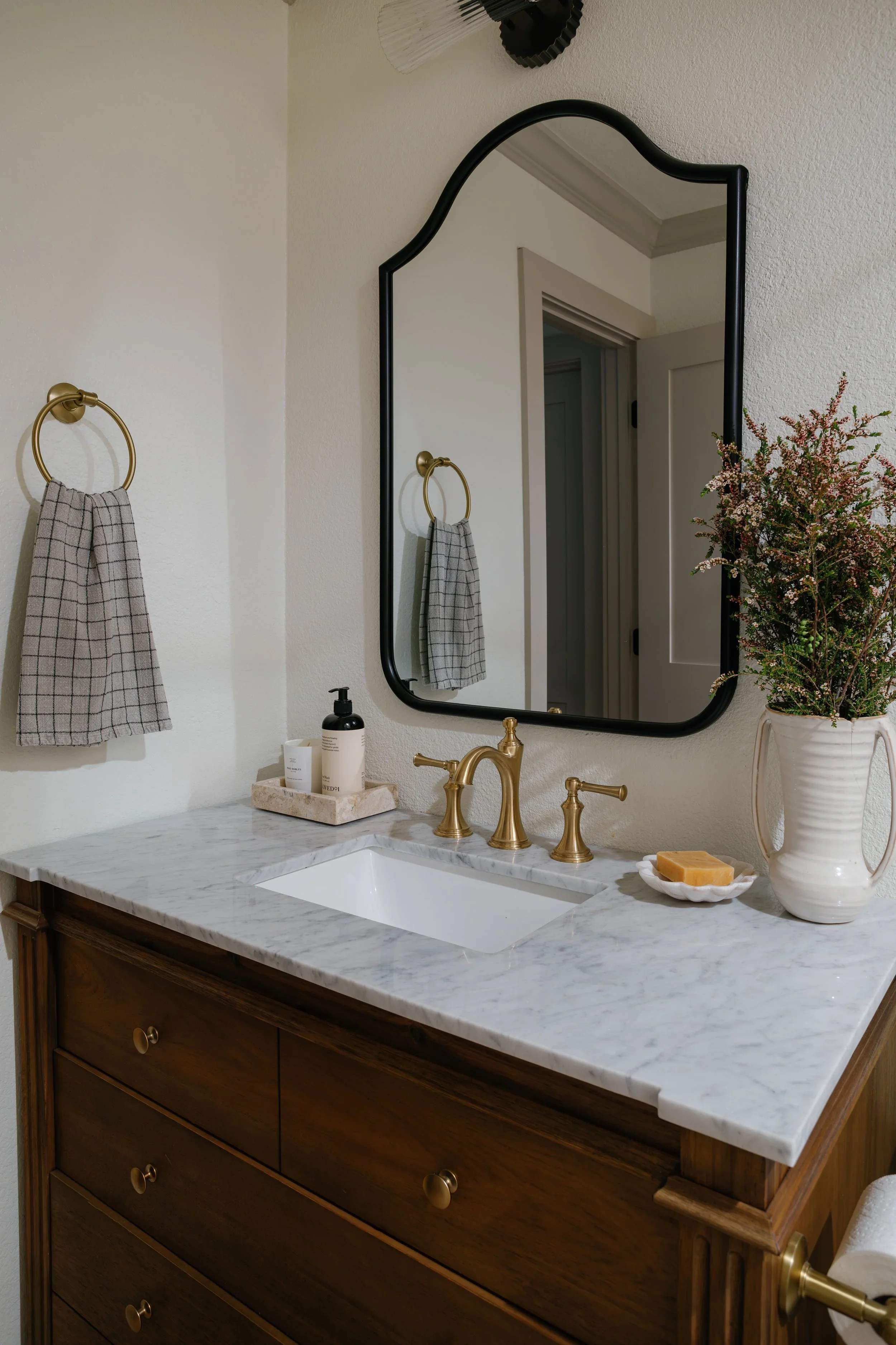 A bathroom vanity with a white marble countertop, a white undermount sink, and a brass faucet. On the countertop, there are bottles of skincare products, a soap bar on a white dish, and a large white vase with pink and green flowers. There is a mirro