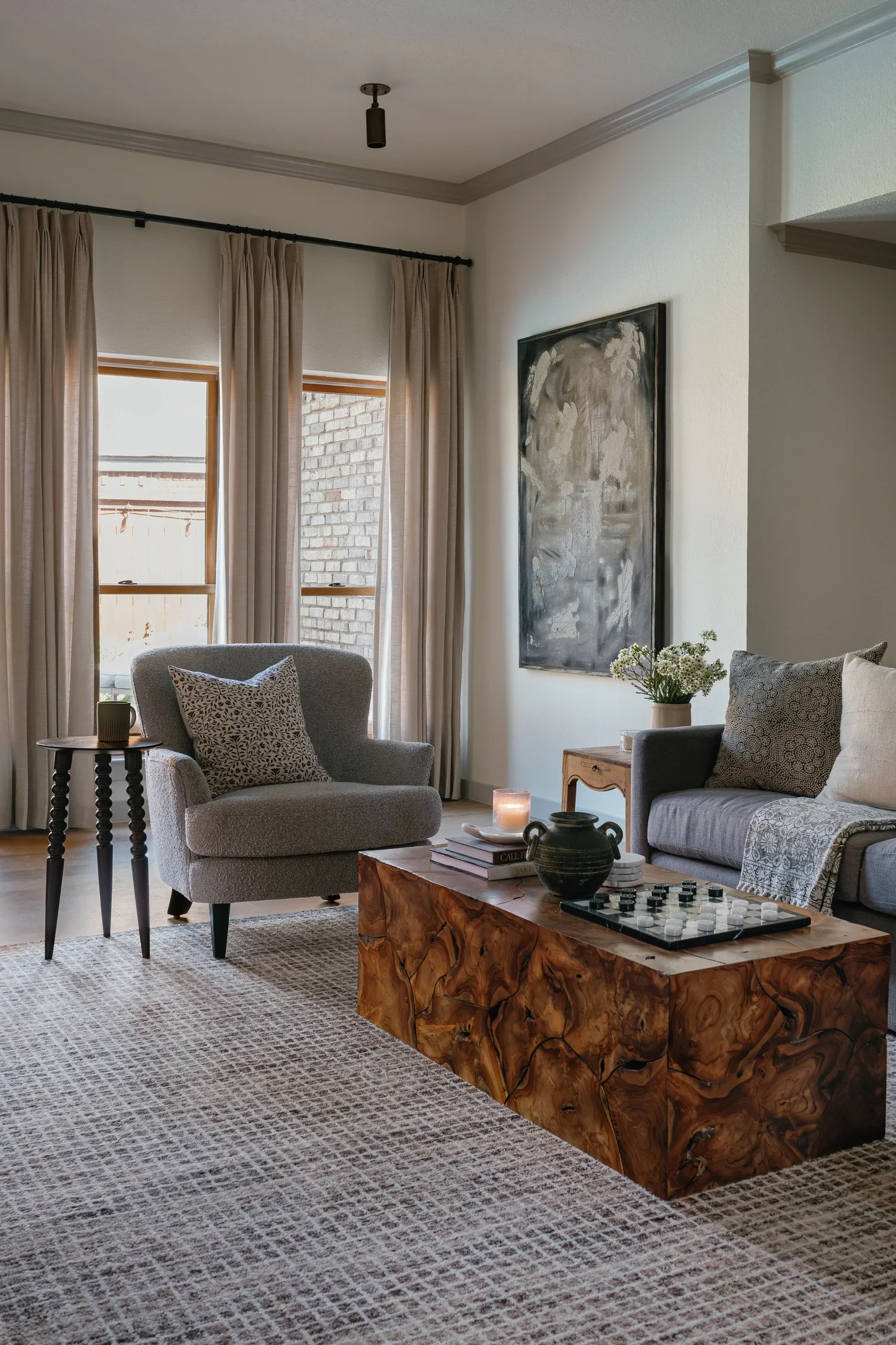 Living room with beige curtains, gray armchair with patterned pillow, abstract wall art, small side table with a green vase, a gray sofa with patterned pillows, a wooden coffee table with a chess set, candlestick, books, and decorative jar, a textured beige rug, and a large window with brick wall outside.
