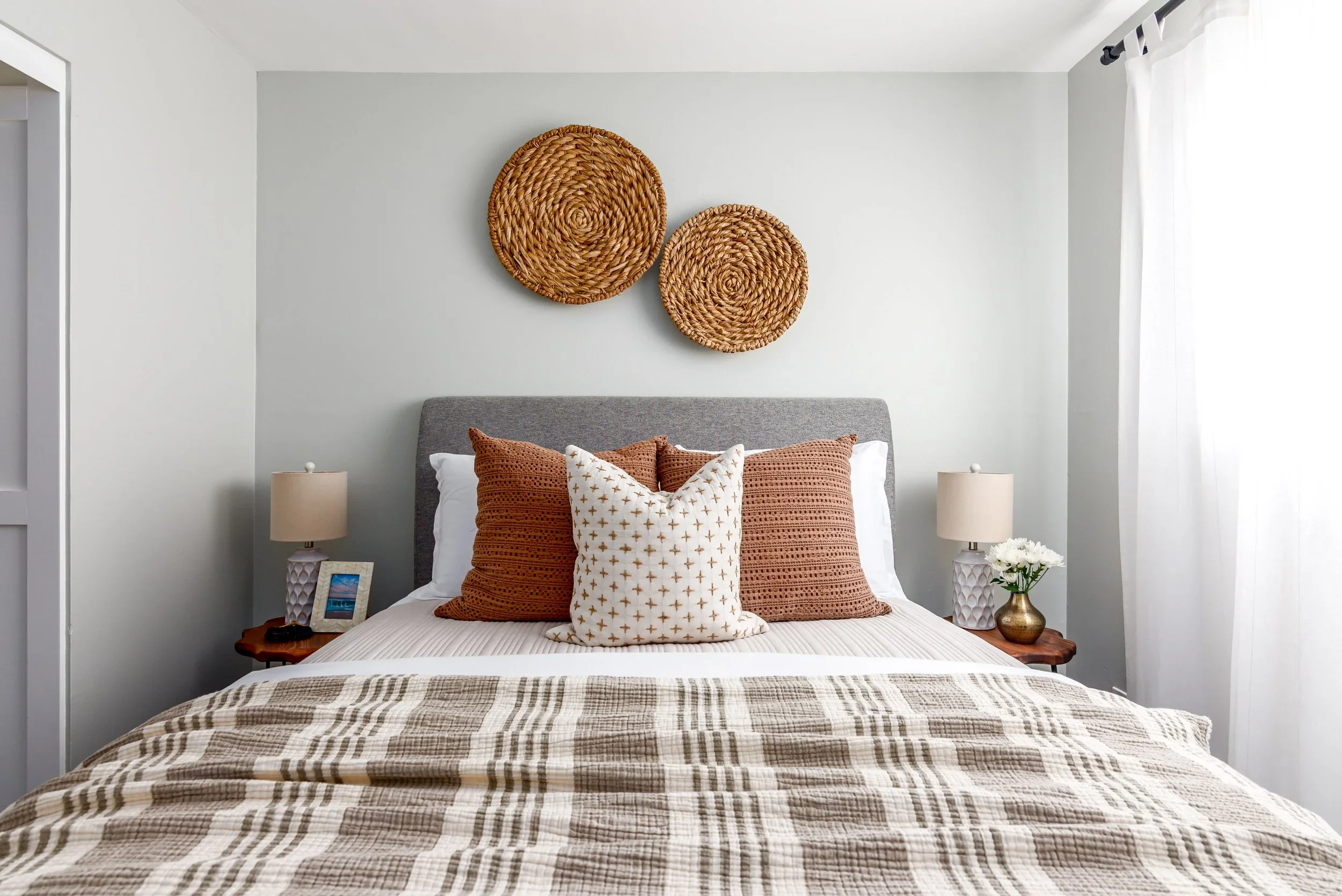 A neatly made bed with a grey upholstered headboard, four decorative pillows in shades of brown and white, and a bedspread with a plaid pattern. On each side of the bed are matching white lampshades and bedside tables, with a small picture frame and 