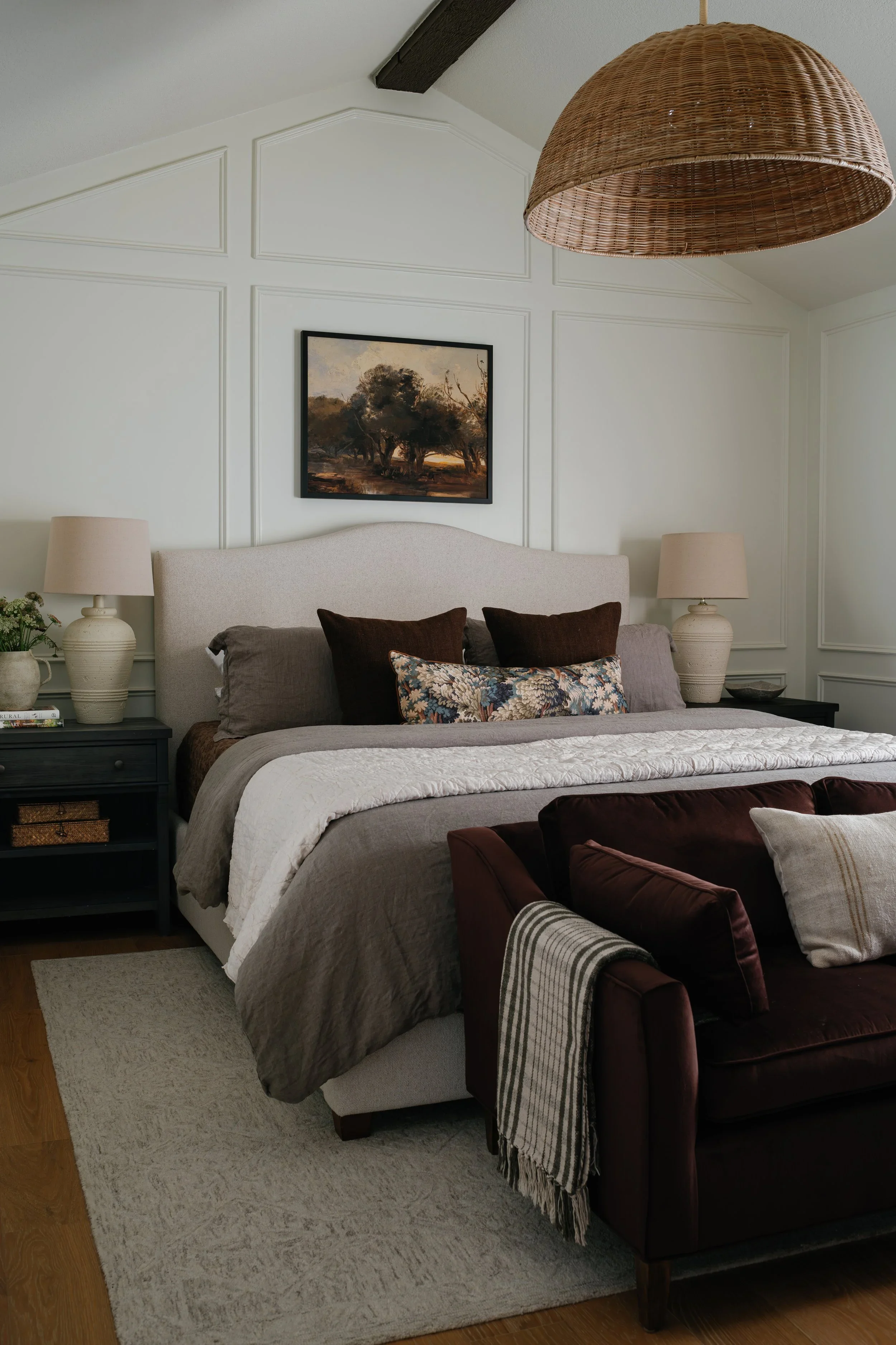 A bedroom with a bed, two nightstands with lamps, a painting of trees on the wall, and a chandelier hanging from the ceiling.