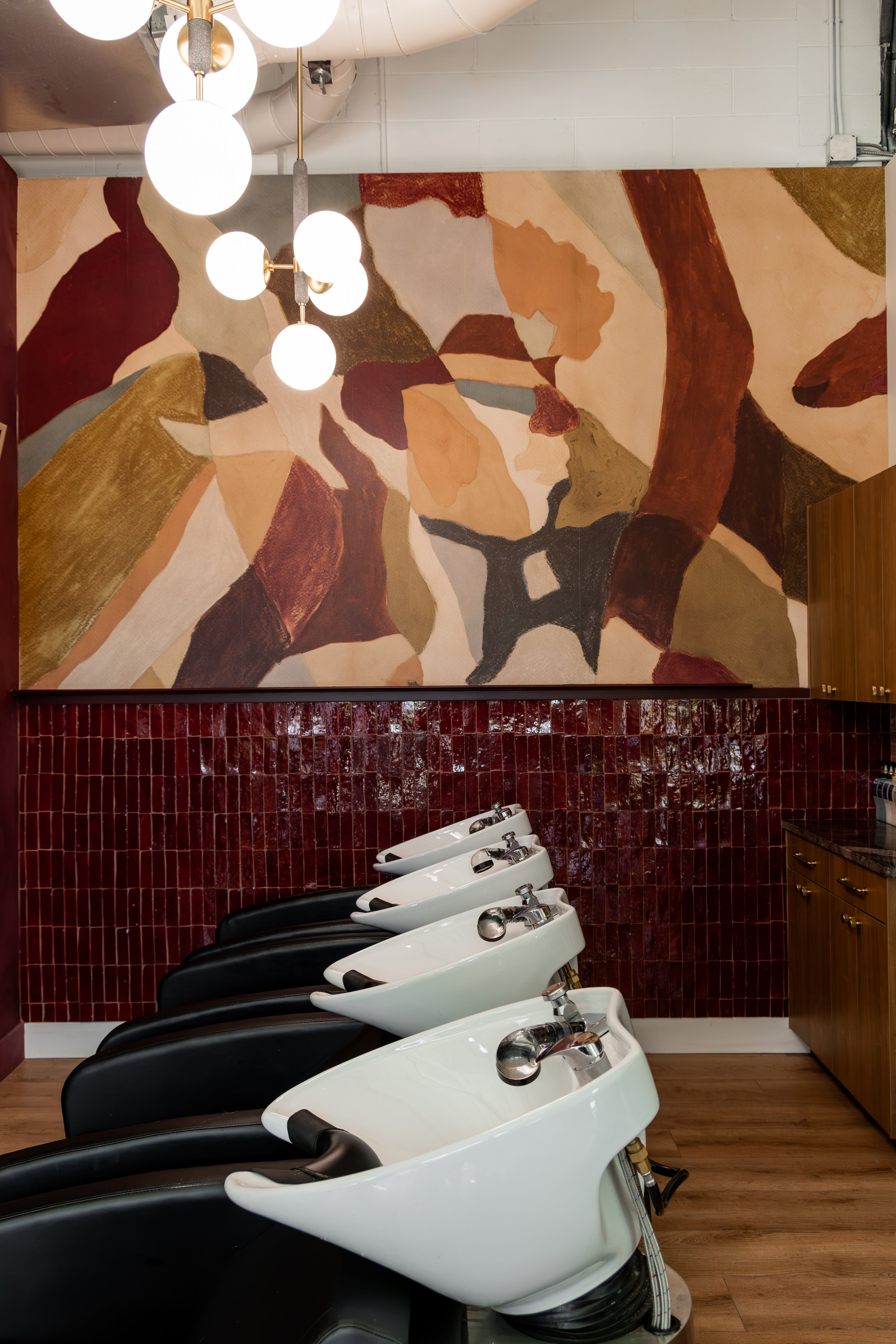A row of black and white salon shampoo stations with sinks and faucets, set against a red tiled wall with an abstract wall art painting above, in a hair salon.