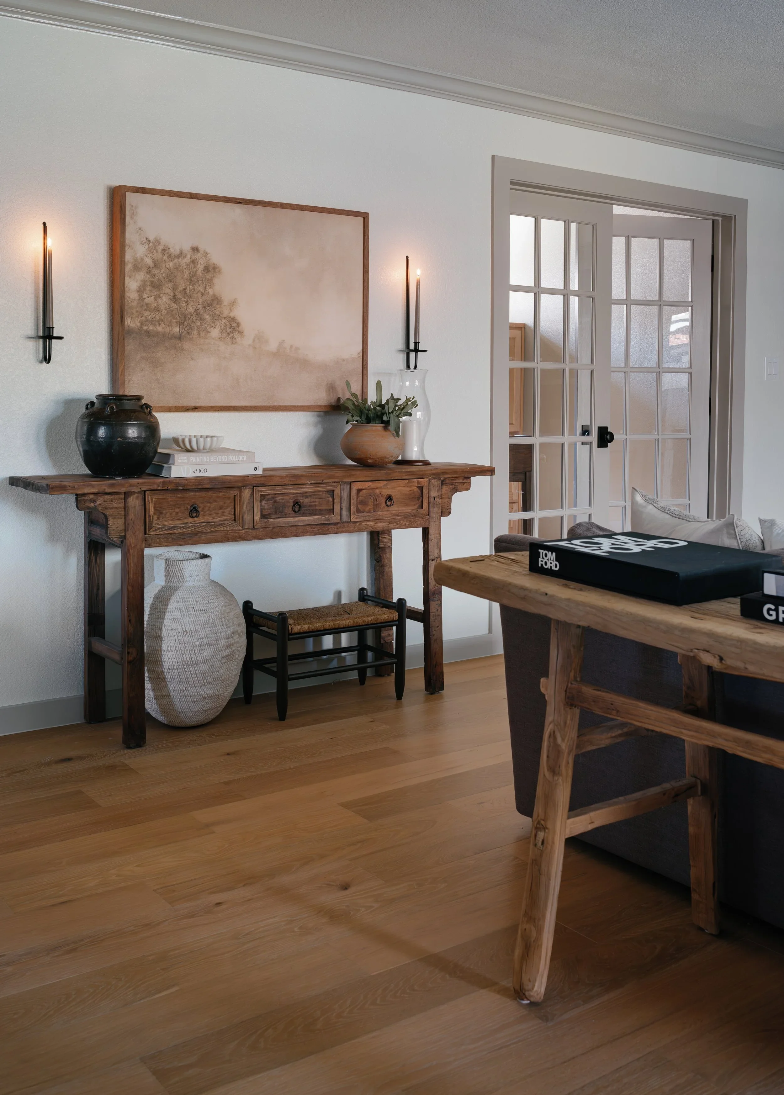 Living room corner with a wooden console table, beige wall with a landscape painting, black vase, books, and a potted plant. A fabric lamp hangs beneath the table, and a black chair is partially visible on the right. Wooden flooring and a glass-paned