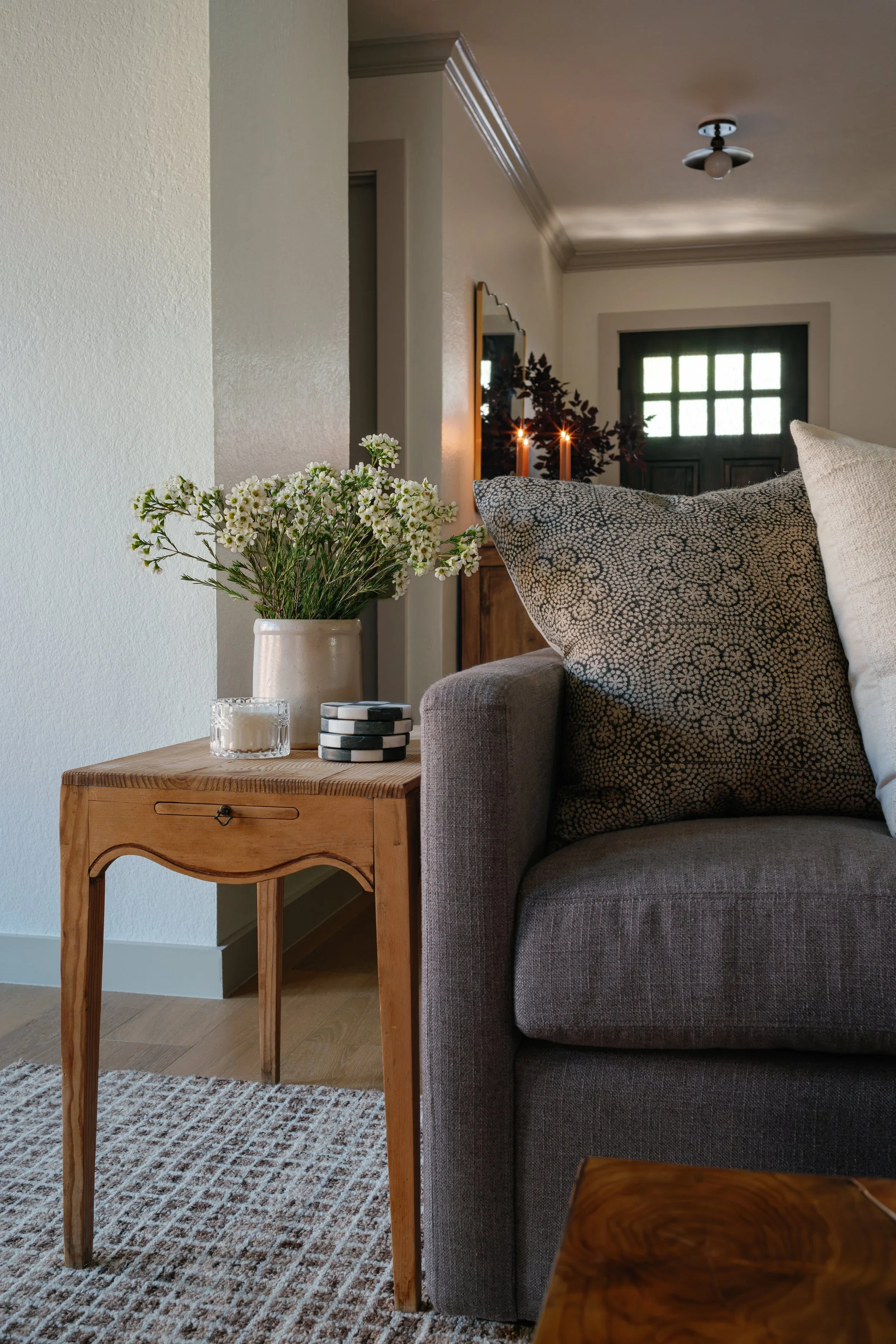 A living room scene with a gray sofa, patterned pillows, a wooden side table with a small vase of white flowers, candles, and a decor piece. In the background, there are candles, a mirror, and a window with green framing.