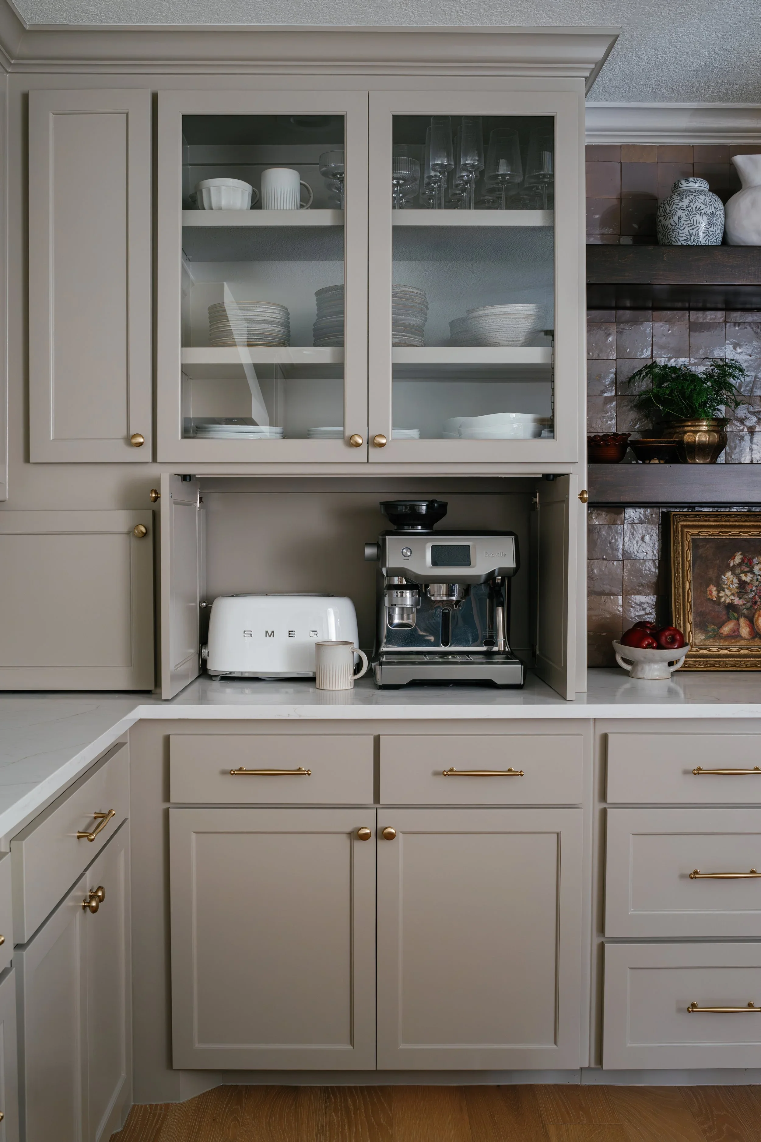 A cream-colored kitchen cabinet with glass doors displaying dishes and glassware, below a coffee machine and toaster on the counter, with a potted plant and decorative jars on a dark wooden shelf to the right.