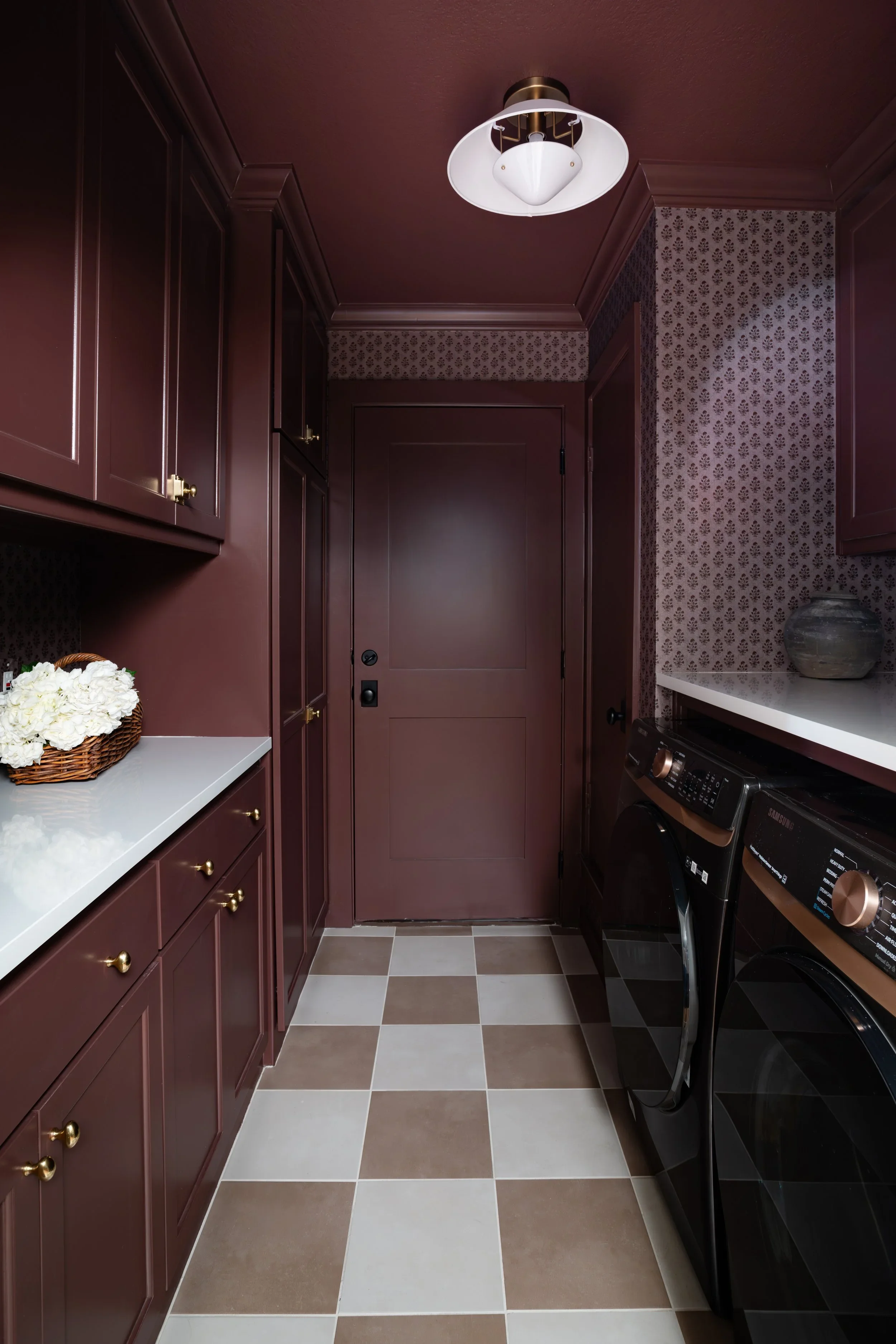 A narrow laundry room with dark red cabinets and gold knobs on the left side, a white countertop with a basket of white flowers, dark red walls with patterned wallpaper, a closed door at the end, and black washing machines on the right side. The floor has a checkered pattern with beige and white tiles.