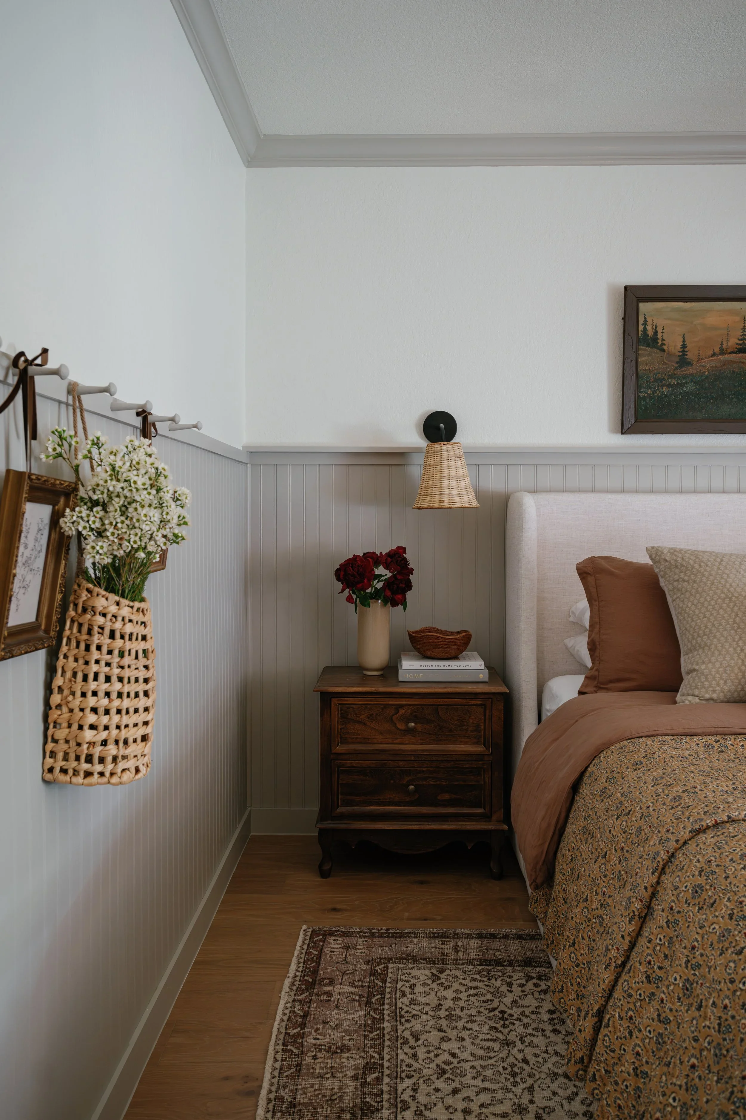 A cozy bedroom corner with a wooden nightstand, beige upholstered headboard, and a bed with brown and patterned bedding. There is a basket with white flowers hanging on the wall, a vase with red flowers on the nightstand, and a framed landscape paint