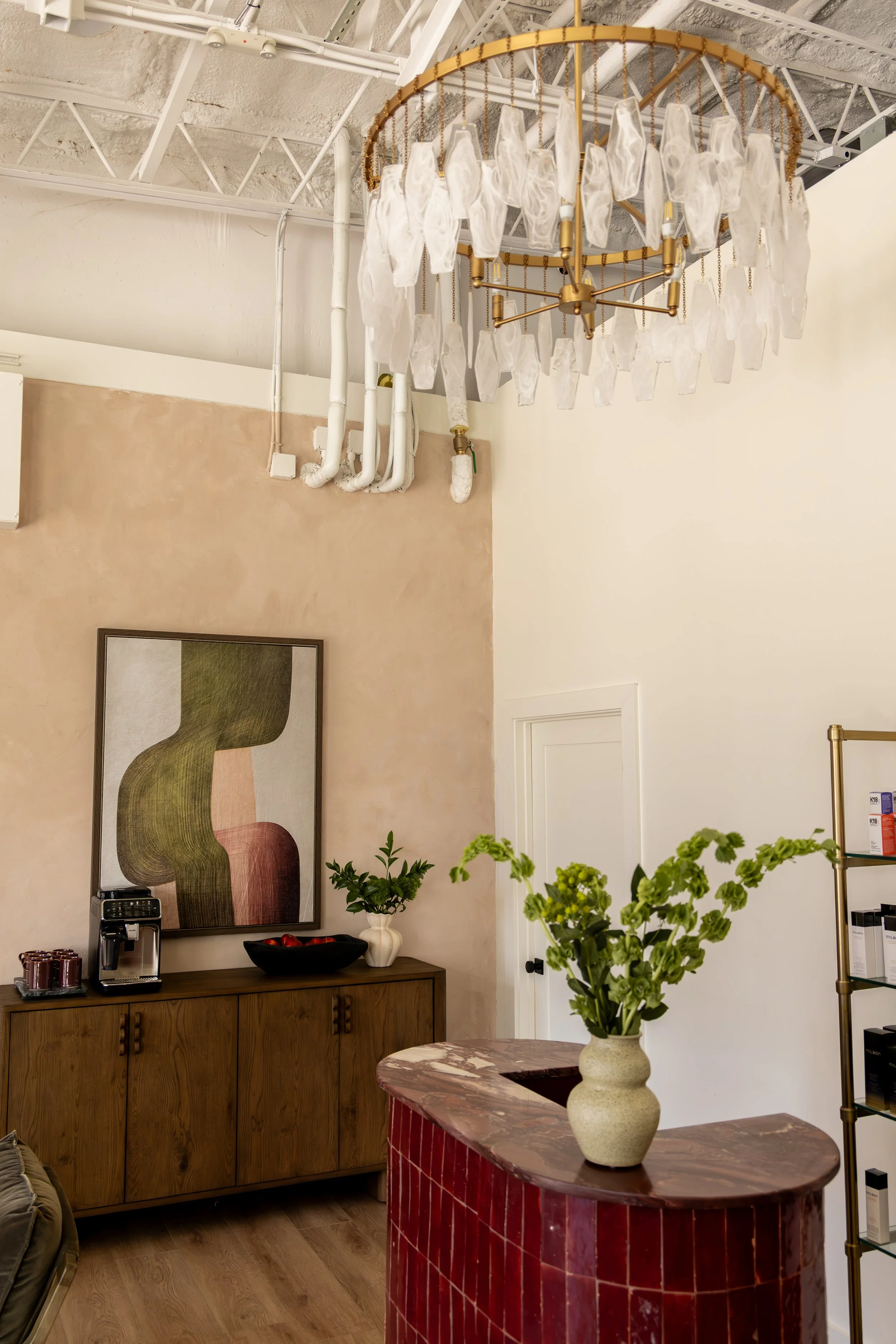 A room with a chandelier hanging from a ceiling, a wooden sideboard with a coffee machine, bowls, and a potted plant, and a painting on the wall. A marble-topped counter with a potted plant is in the foreground.