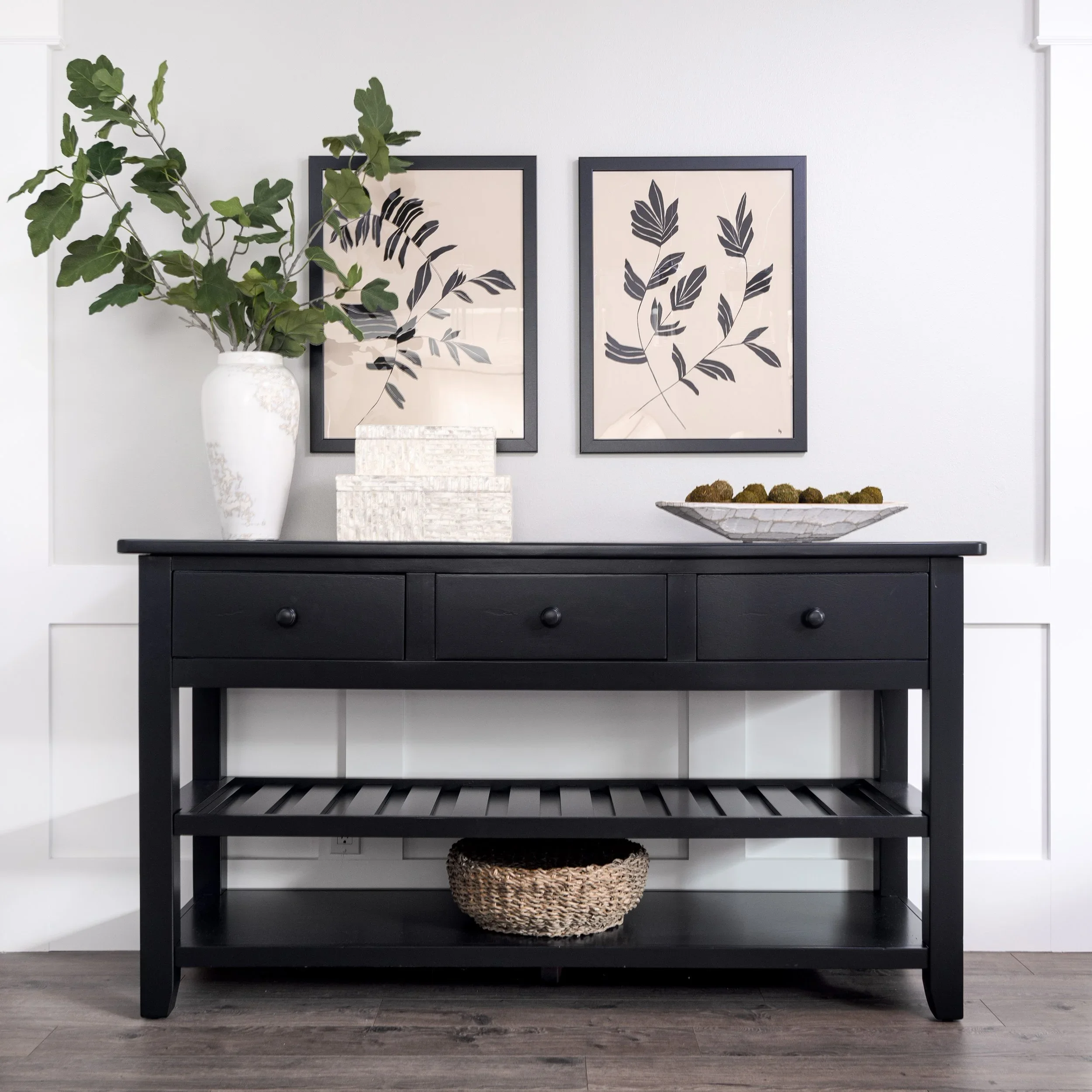 A black wooden console table with two drawers under a dark tabletop against a white wall. Two framed art prints with botanical leaf illustrations in black are hung above. On the left, a white vase with green leafy branches. On the right, a woven bask