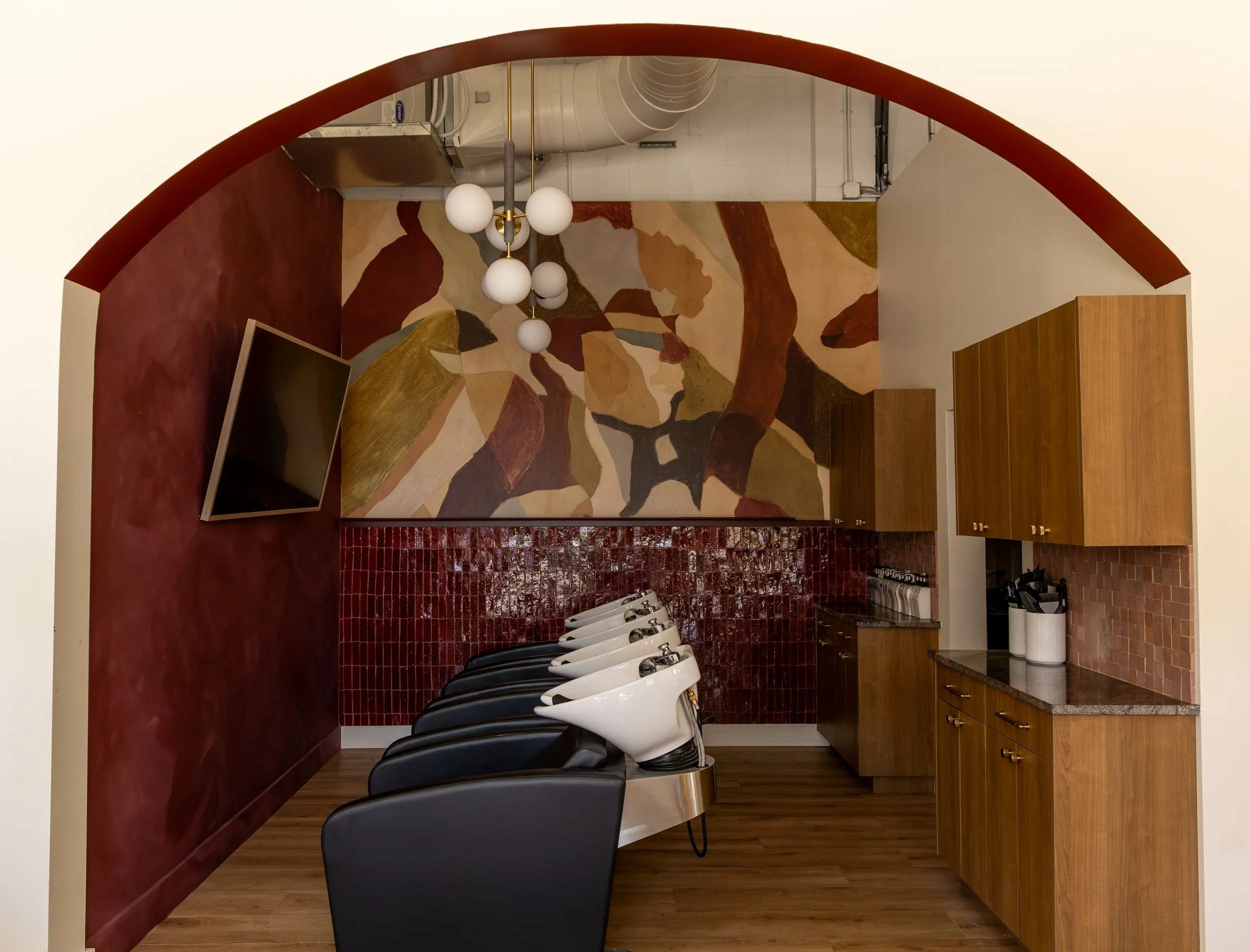 Interior view of a salon or barber shop with a row of white sinks and black chairs, wooden cabinets, red tiled wall, a large abstract painting, a chandelier with globe lights, a wall-mounted television, and a kitchen area.