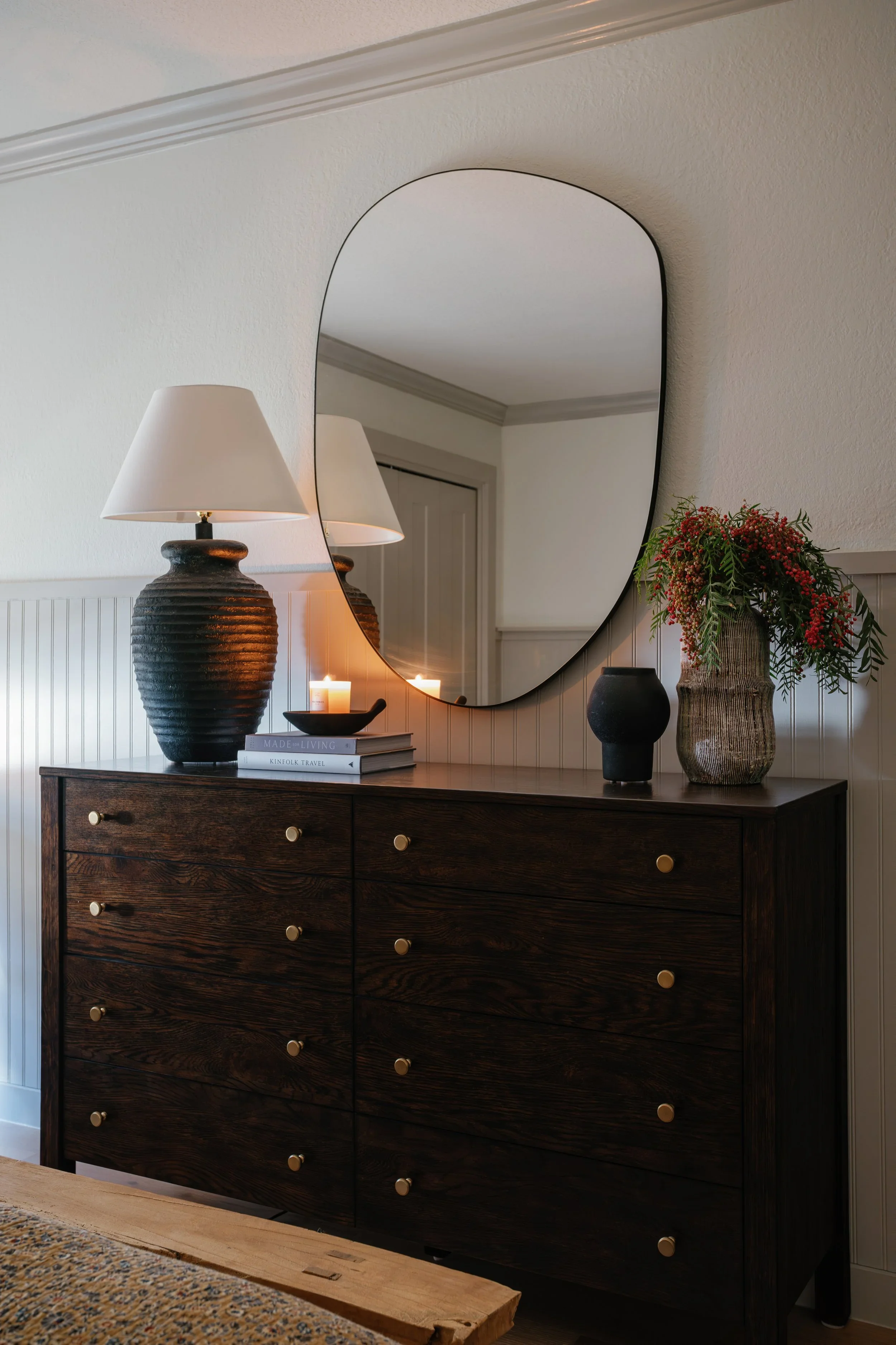 A dark wooden dresser with twelve drawers, each with a round brass knob, placed against a white wall with beadboard paneling. On top of the dresser are two black textured lamps with white shades, a small black decorative bowl, a stack of books, a lit