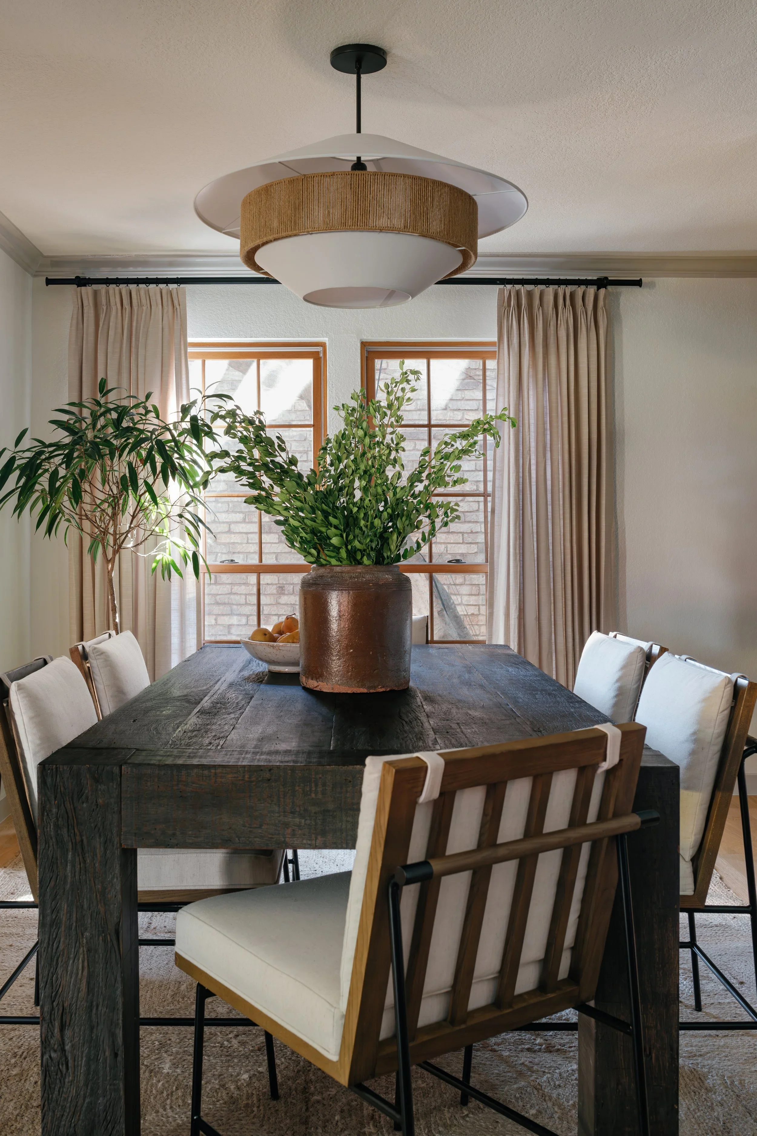 A dining room with six chairs around a dark rustic wooden table, a large potted plant in a brown vase in the center, two windows with beige curtains, a bowl of fruit, and a modern chandelier hanging from the ceiling.