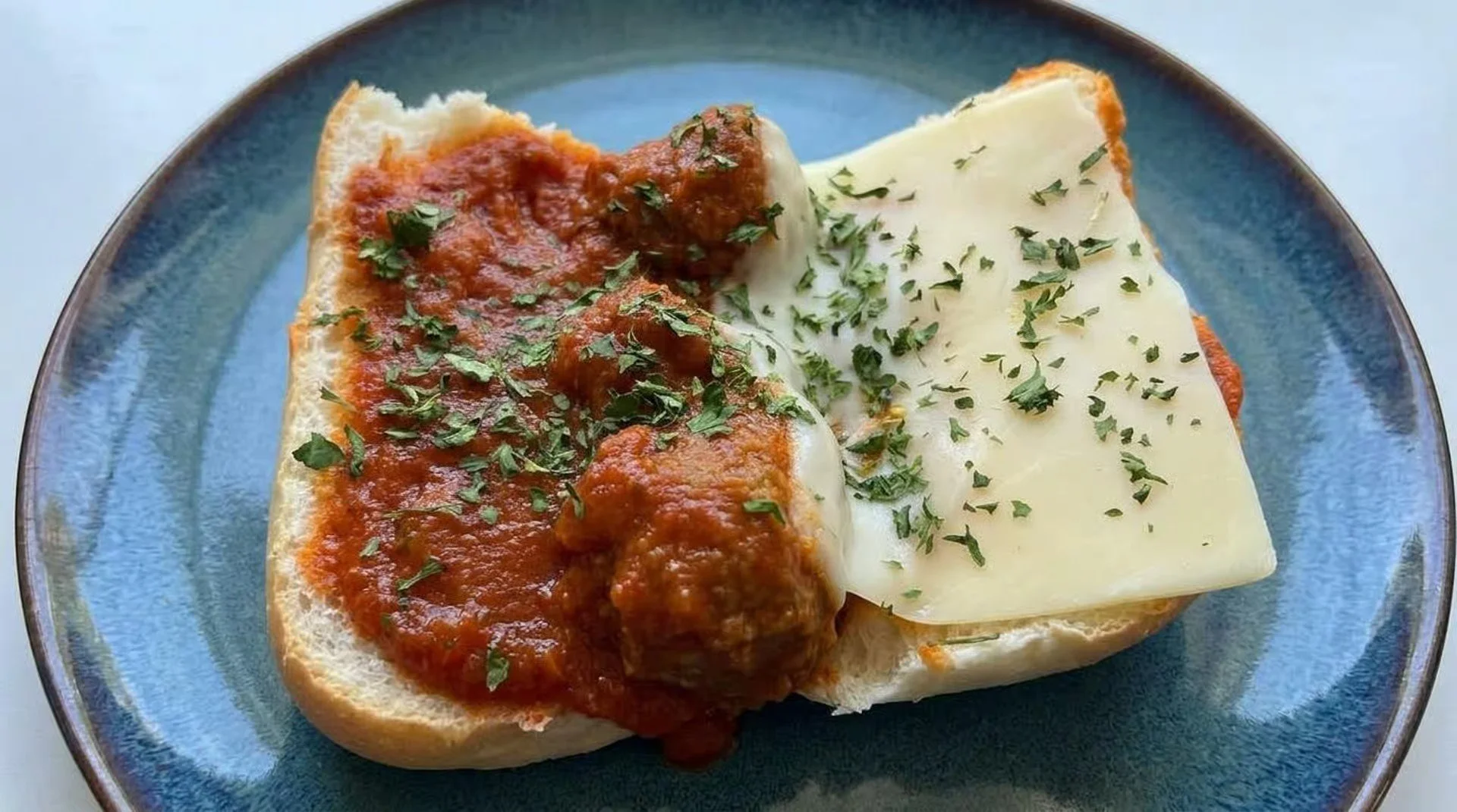 Open-faced sandwich with meatball, tomato sauce, and cheese on bread, garnished with chopped herbs, served on a blue plate.