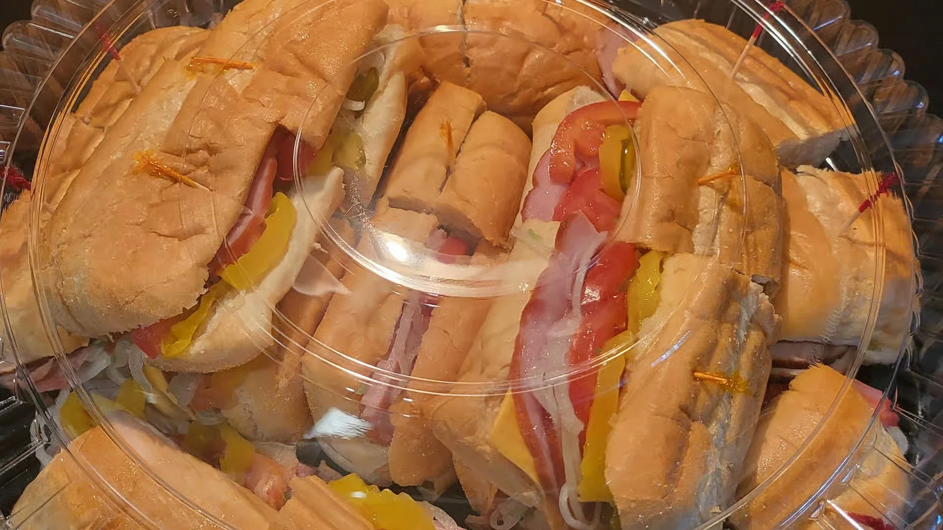 A plastic container filled with various sub sandwiches containing meats, cheese, lettuce, tomato, and pickles.