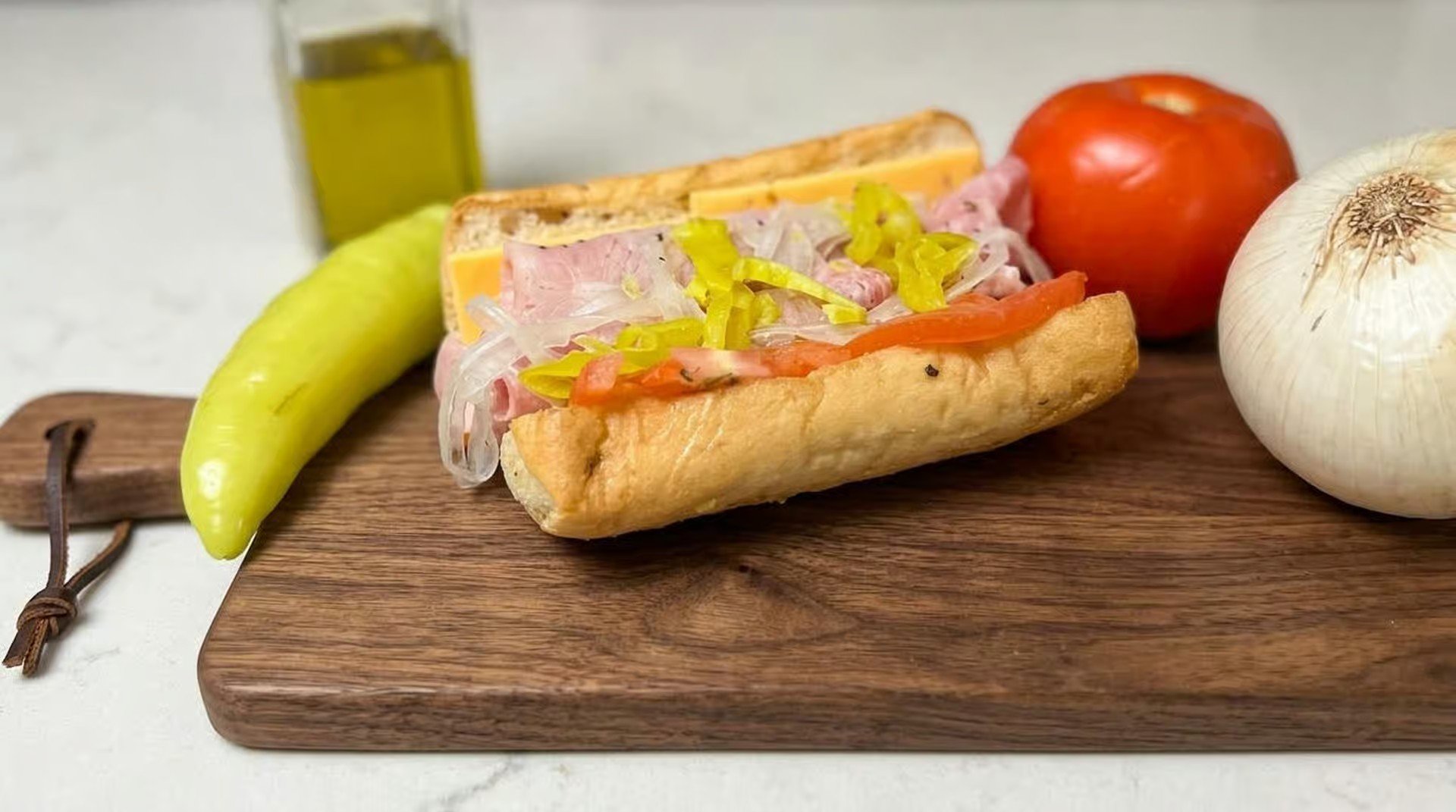 Hot dog with yellow mustard, onions, and tomatoes on a bun, placed on a wooden cutting board. Nearby are a banana, a red apple, and a whole bulb of garlic, with a bottle of olive oil in the background.