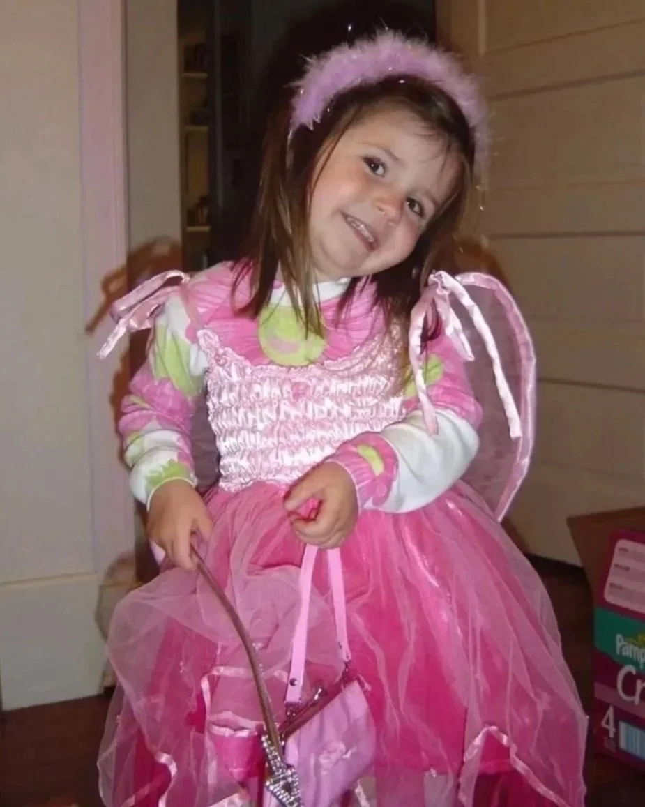 A young girl dressed in a pink princess costume with a tutu, fairy wings, and a fluffy headband, sitting on a chair indoors.