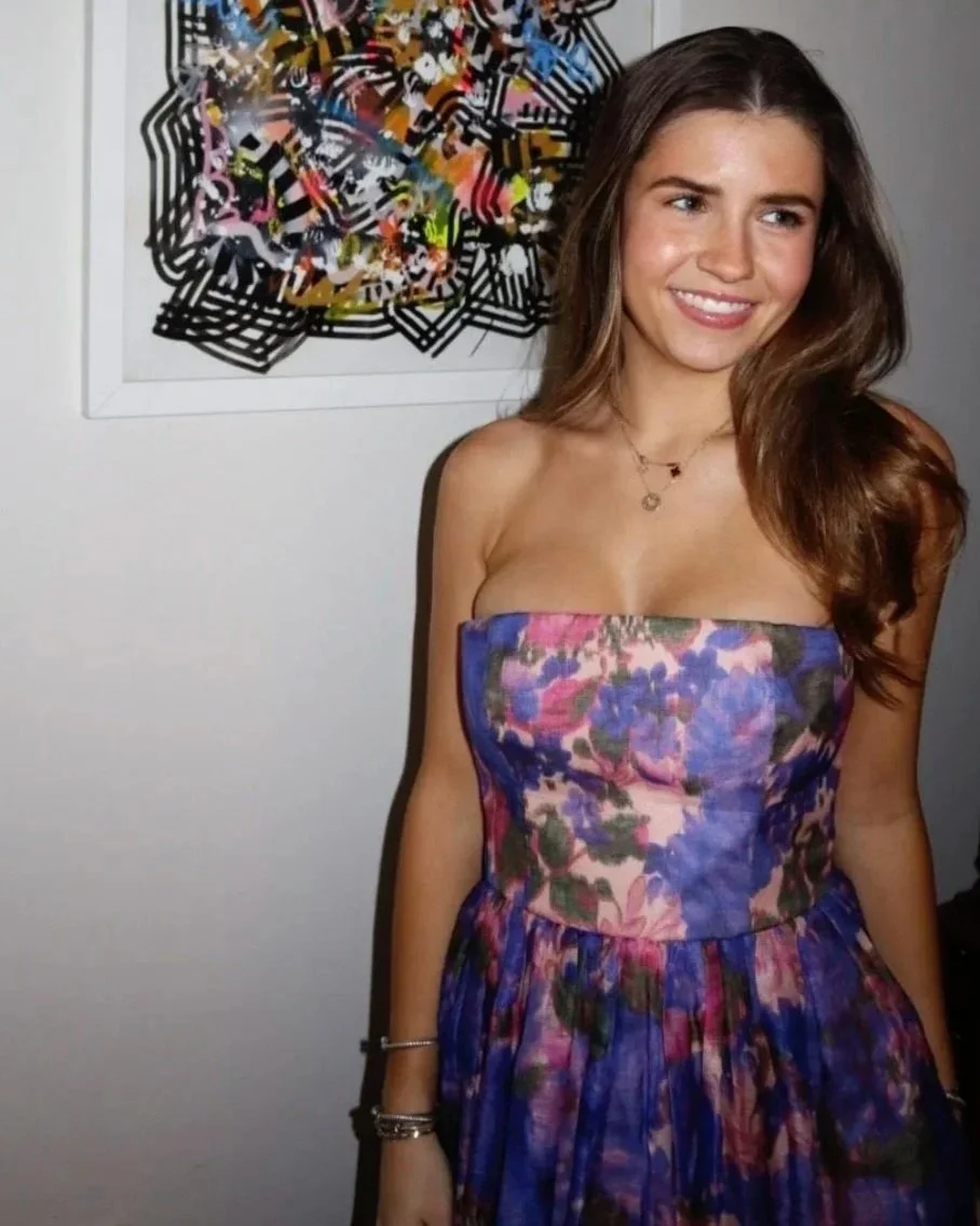 A young woman with long brown hair smiling, wearing a colorful strapless floral dress, standing in front of abstract artwork on a white wall.