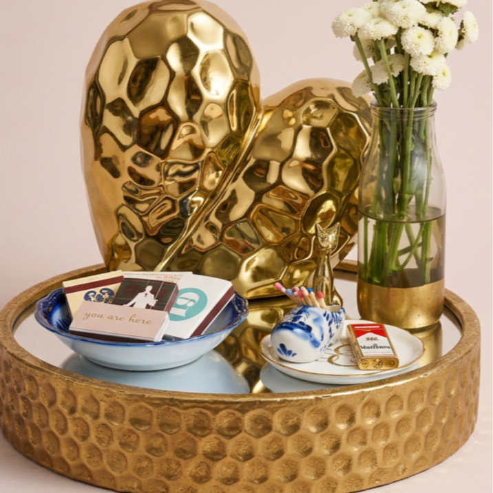 Decorative tray with gold pineapple and leaf sculptures, a glass vase with white flowers, small bowls with stationery and a pack of cigarettes.