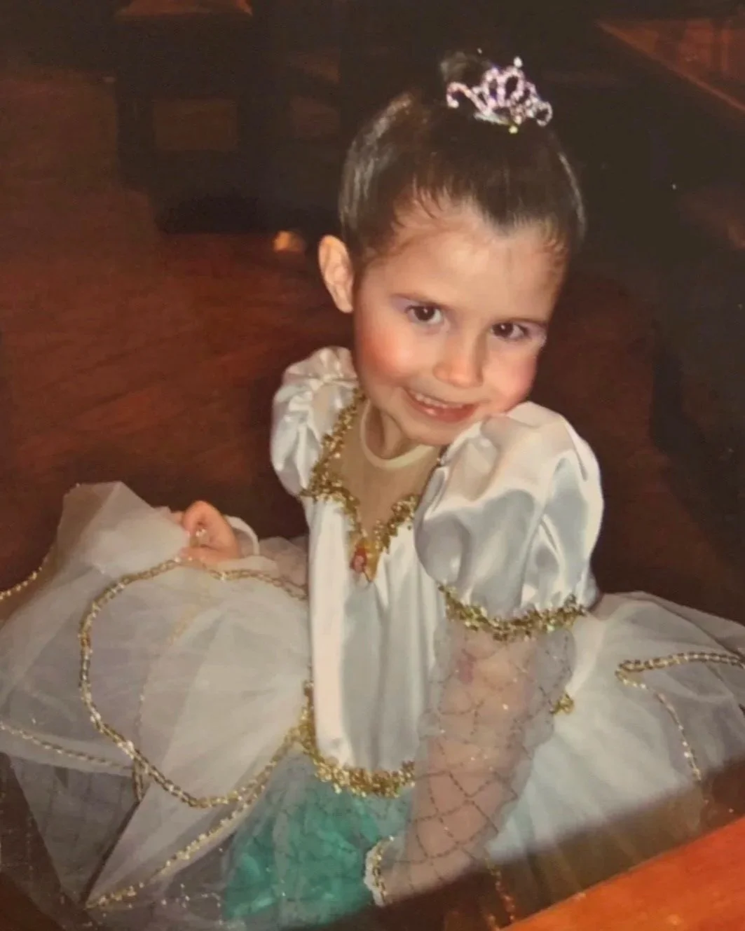 A young girl dressed as a princess, wearing a tiara, shiny white dress with gold accents, and jewelry, smiling at the camera.