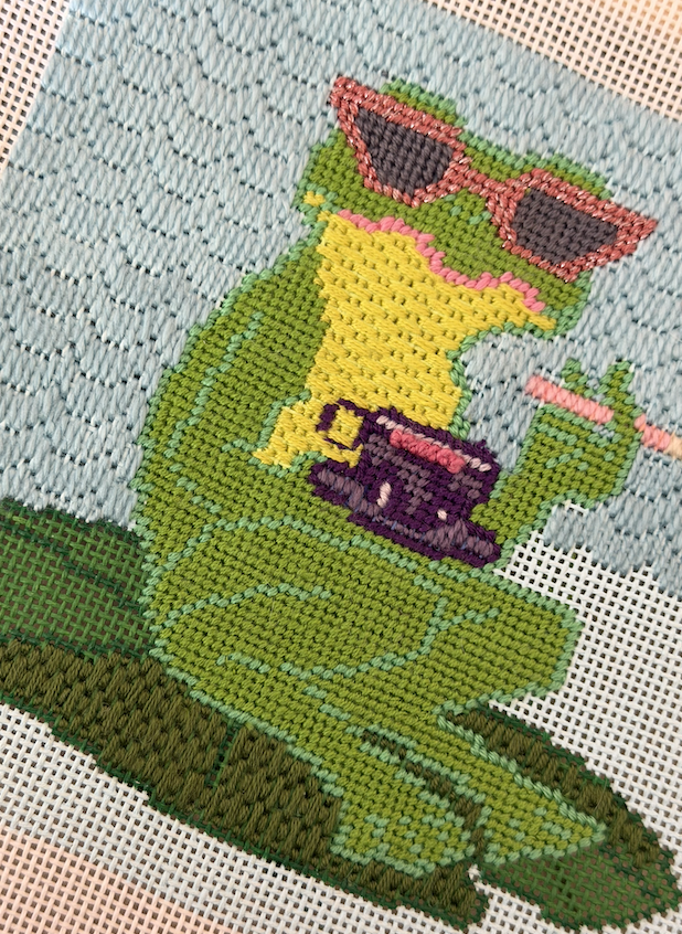Cross-stitch embroidery of a green dinosaur wearing pink sunglasses, holding a purple cup, and sitting on a green patch of grass.