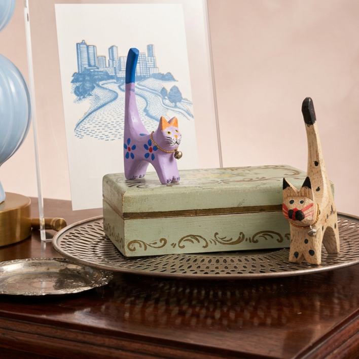 Decorative wooden cat figurines, one painted purple with blue spots and a bell, and the other carved wooden with a long neck and spots, placed on a wooden surface with a decorative tray and a hand-drawn art of a cityscape in the background.