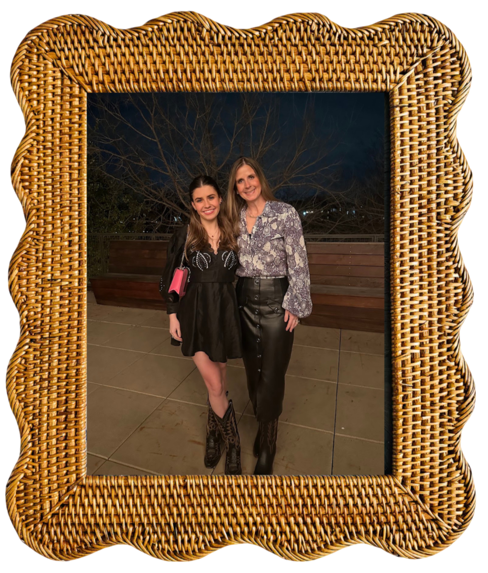 Two women smiling and standing close together during nighttime, framed by a wicker picture frame, on an outdoor terrace with trees in the background.