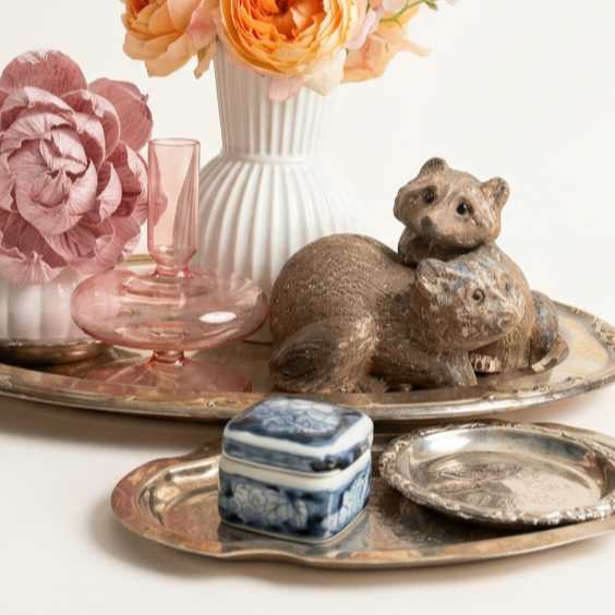 Decorative tray with ceramic animal figurines, pink glassware, a white vase with pink and peach flowers, and metallic plates.