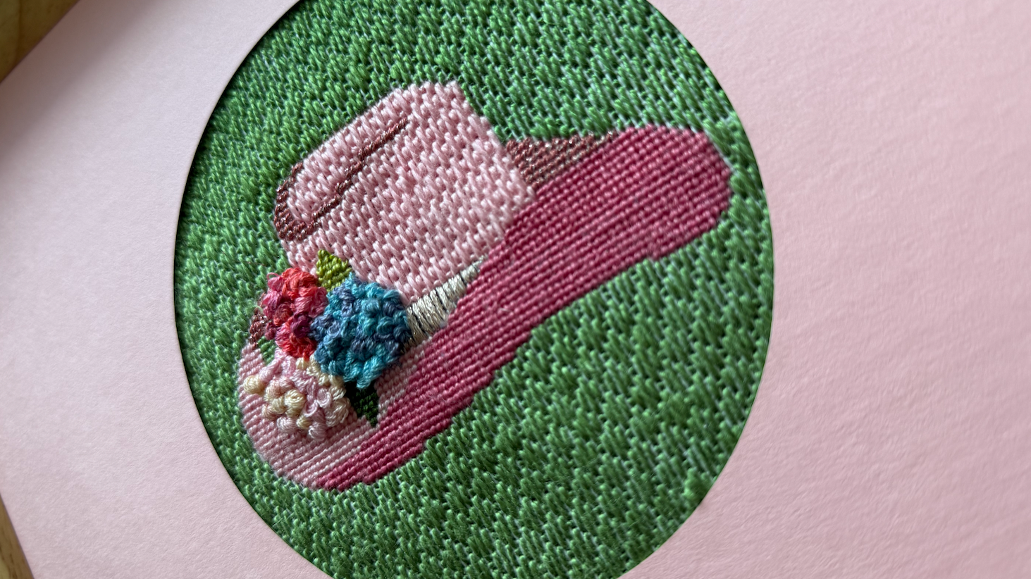 Embroidery of a pink flower with green background and a red, blue, white, and pink flower in the center on pink fabric.
