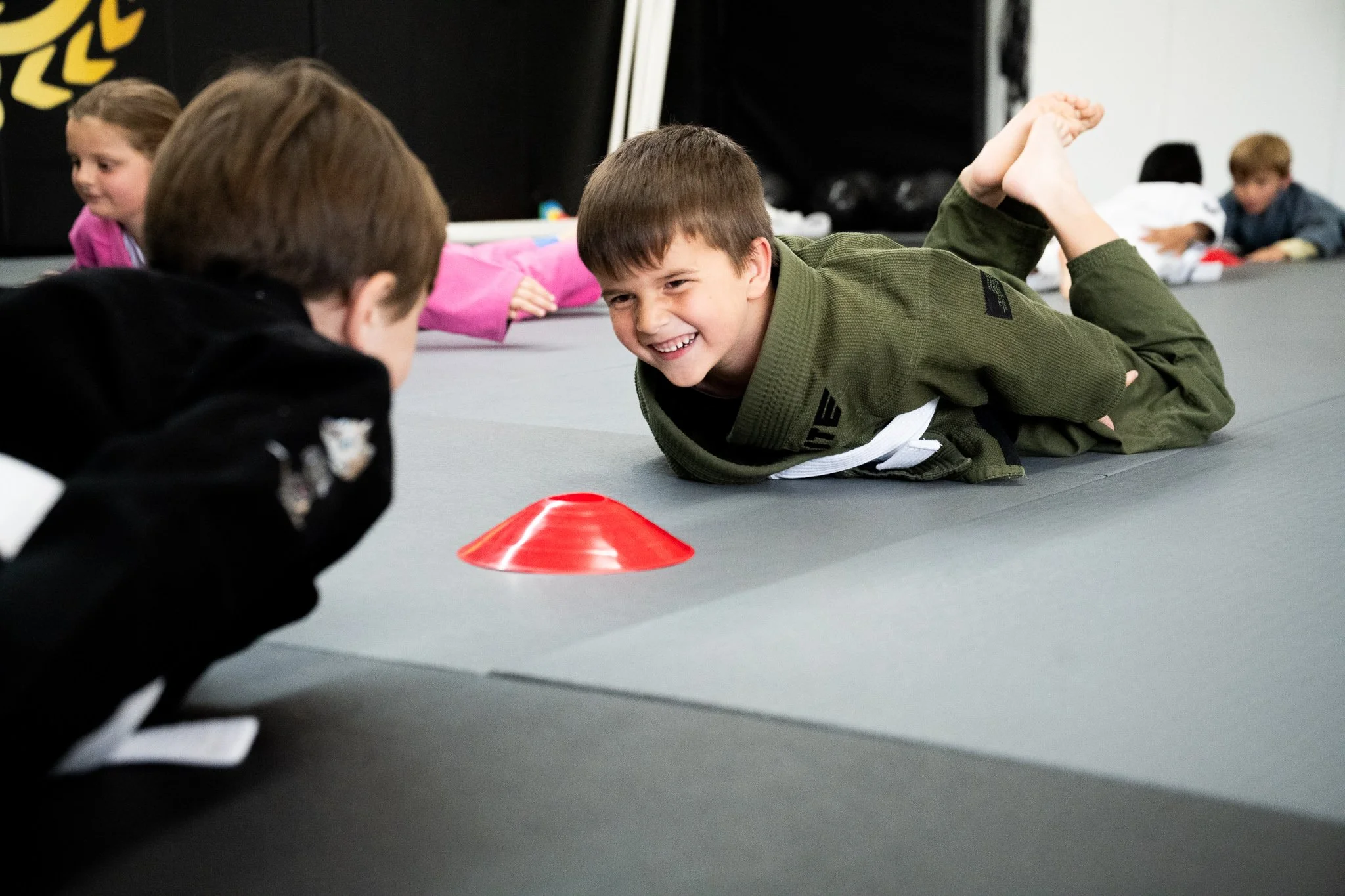 Children practicing martial arts in a gym, lying on a gray mat with small orange traffic cones as markers.