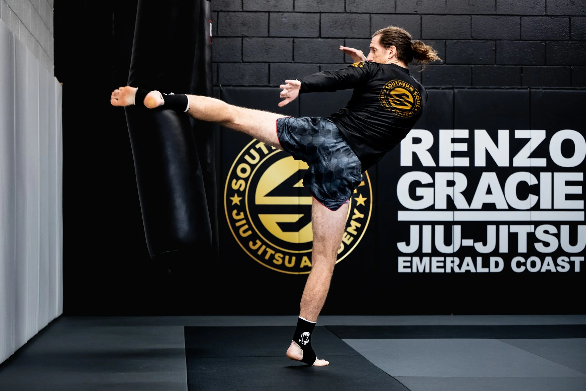 A male martial artist practicing kicking a black padded kick shield held against a wall in a gym. He is wearing black gloves and shorts, and has his hair tied back. The background displays a black wall with logos and text related to martial arts and fitness.