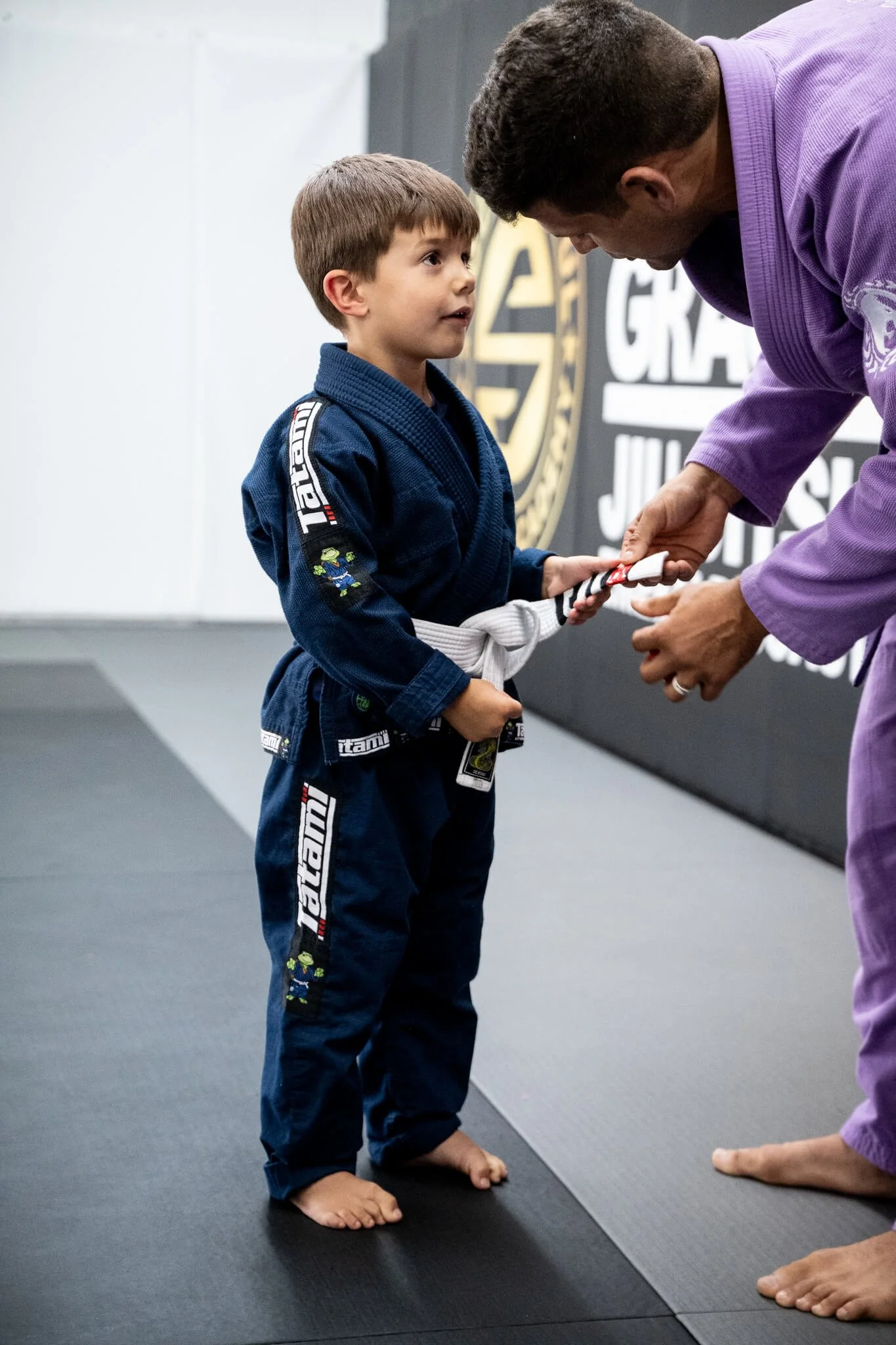 A young boy in a blue martial arts gi with patches and a white belt is receiving his yellow belt in Brazilian Jiu-Jitsu from an instructor wearing a purple gi. They are standing barefoot on a black mat in a martial arts training facility.