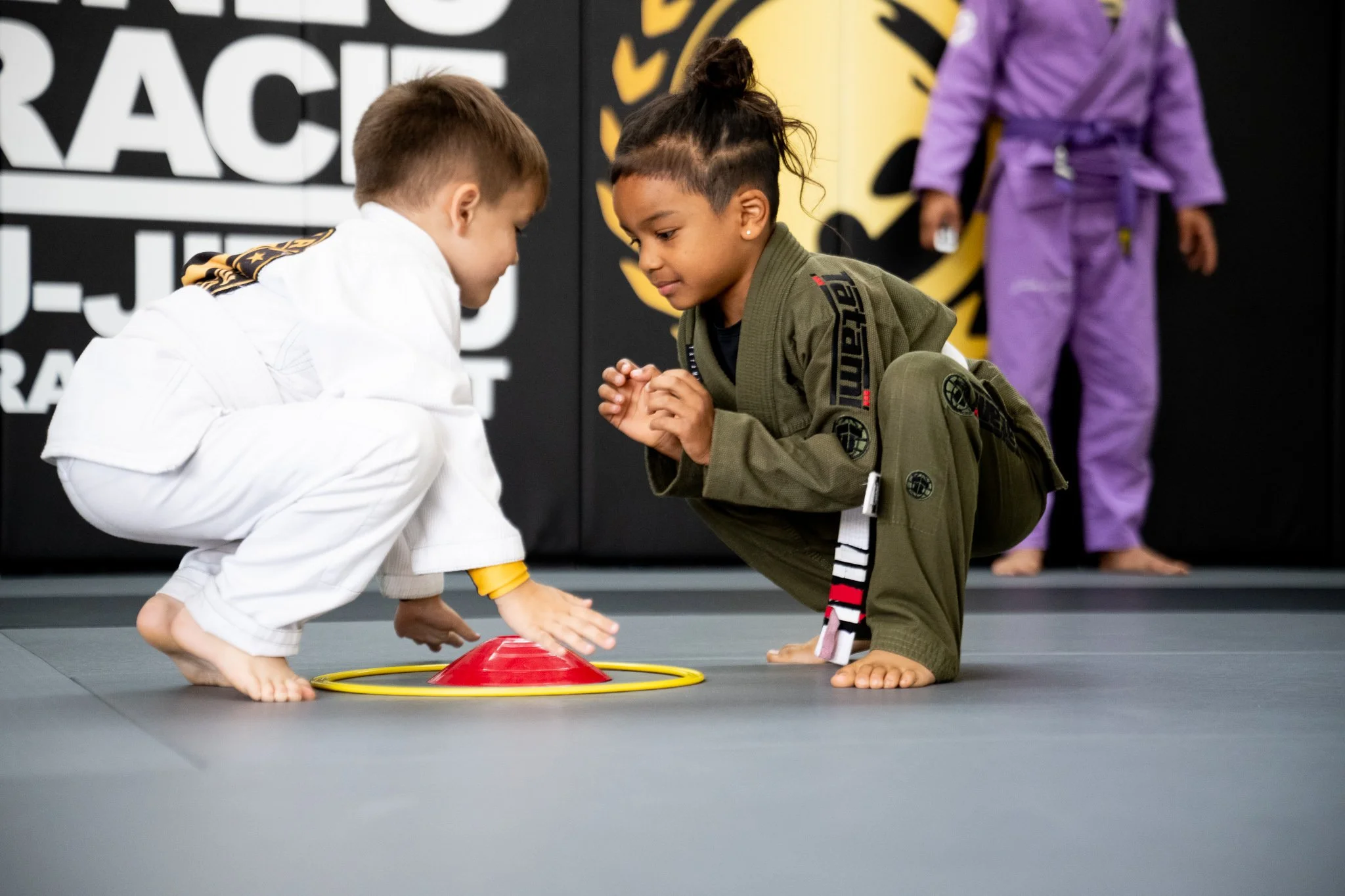 Two children dressed in martial arts gis, one in white and one in green, are kneeling on a martial arts mat and holding each other's hands in a training exercise. There is a red training pad and a yellow ring between them. A third child in a purple gi is partially visible in the background.