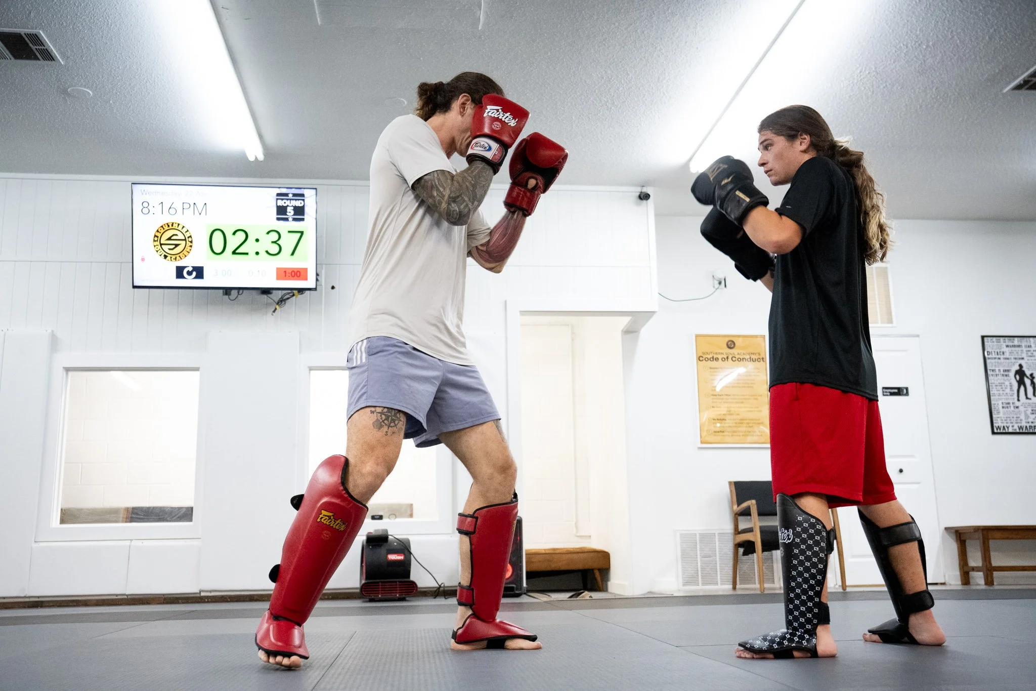 Two women practicing martial arts in a gym, one in red protective gear, the other in black attire with shin guards, facing each other in fighting stances.